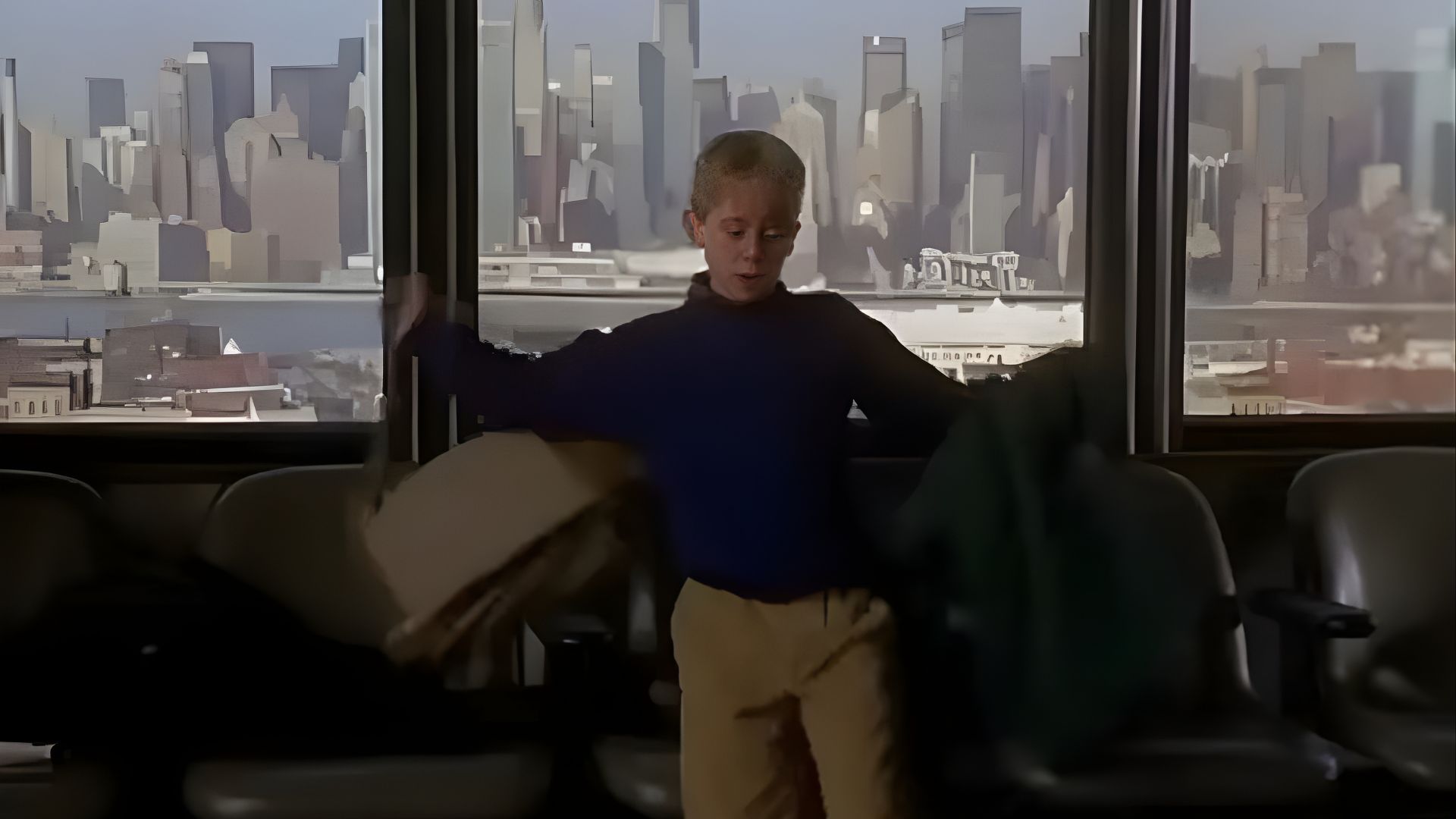 A young boy is positioned in front of a window, with a cityscape visible behind him.