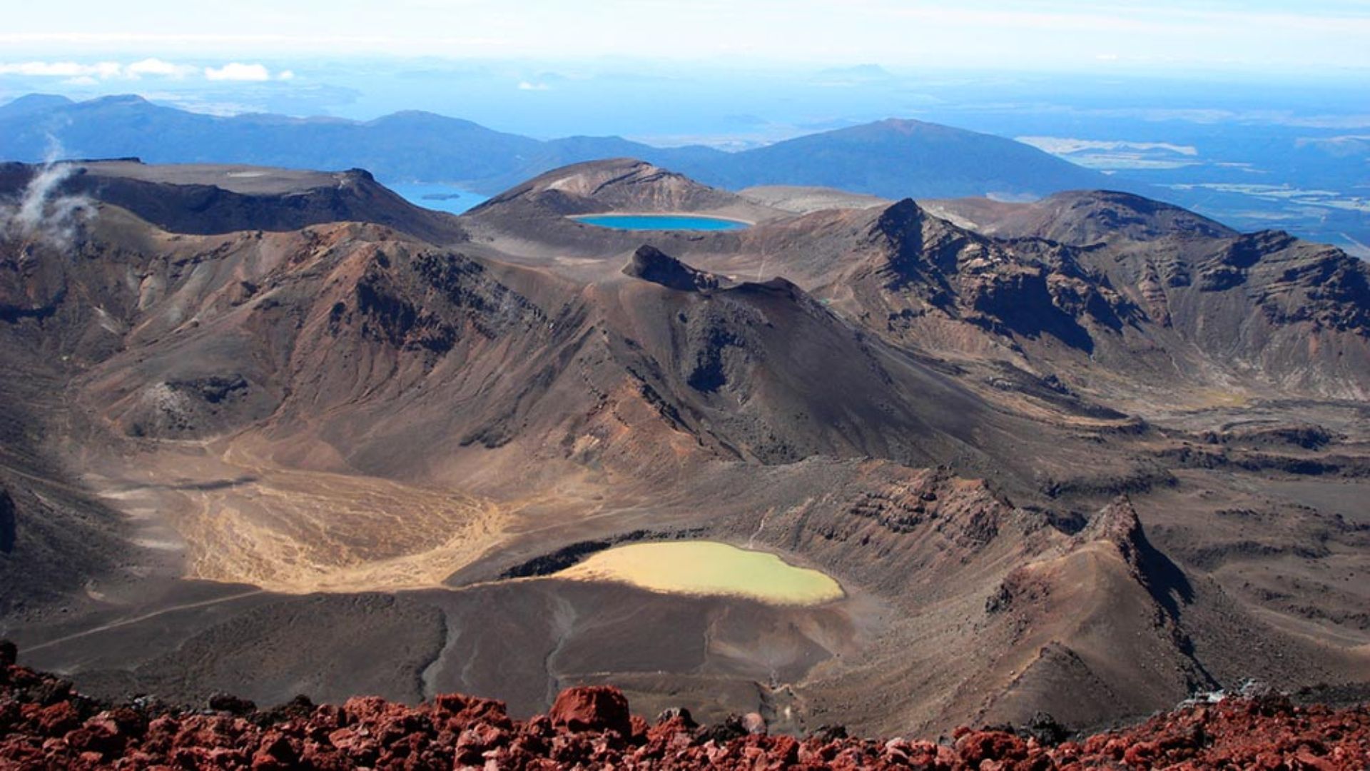 Panoramic view from Mount Tongariro's summit, showcasing volcanic landscapes and distant mountains under a clear blue sky.