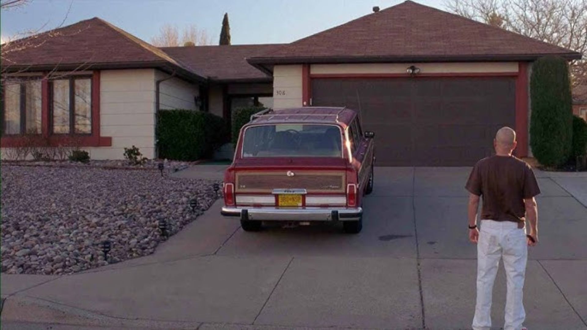 A man stands in front of a house with a car parked in the driveway, showcasing a suburban setting.
