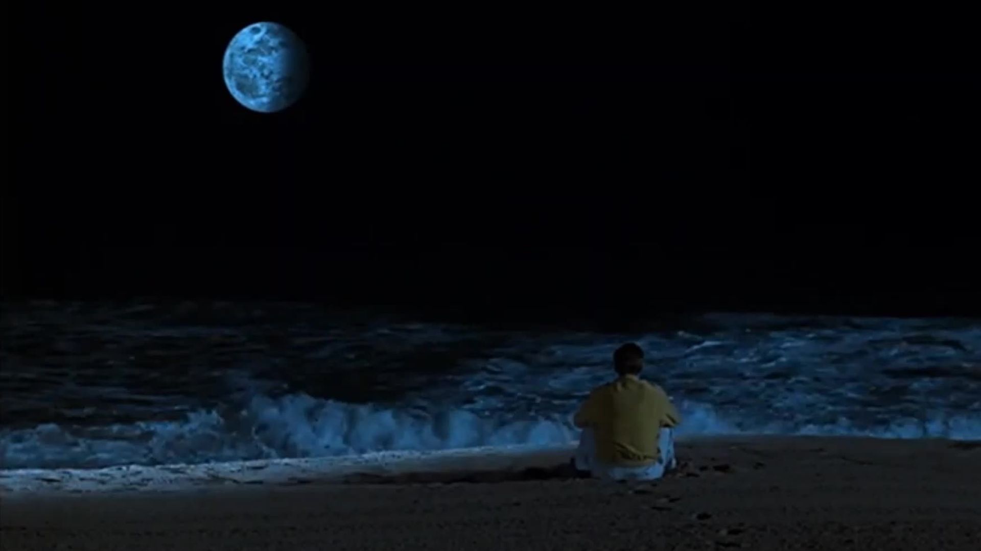 A man sits on the beach, gazing at the moon illuminating the night sky.