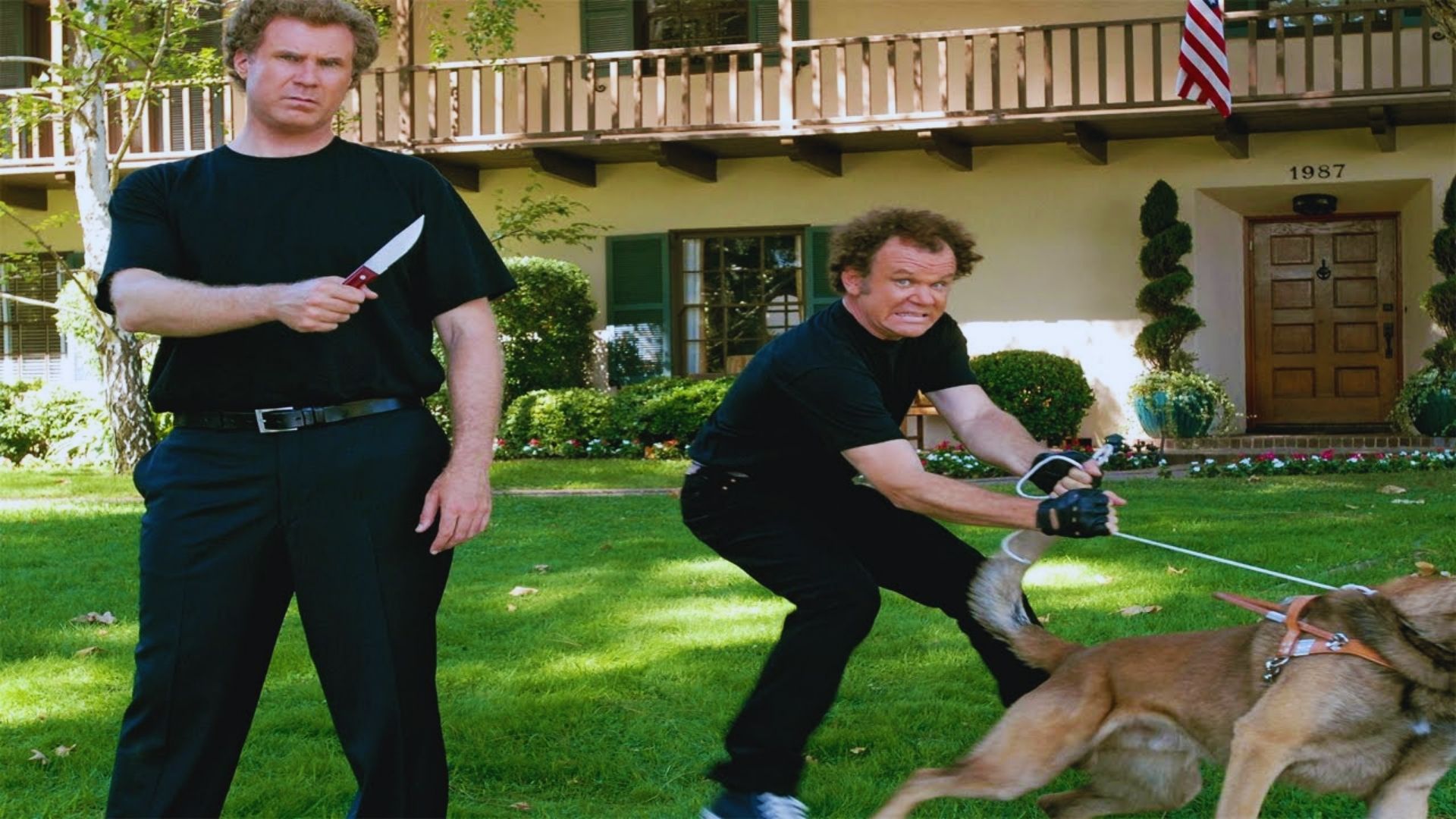 Two men in black attire hold a knife and a dog, standing together in a neutral setting.