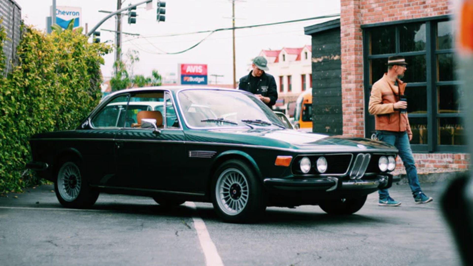 Two men walking down the street beside a green BMW parked on the side.