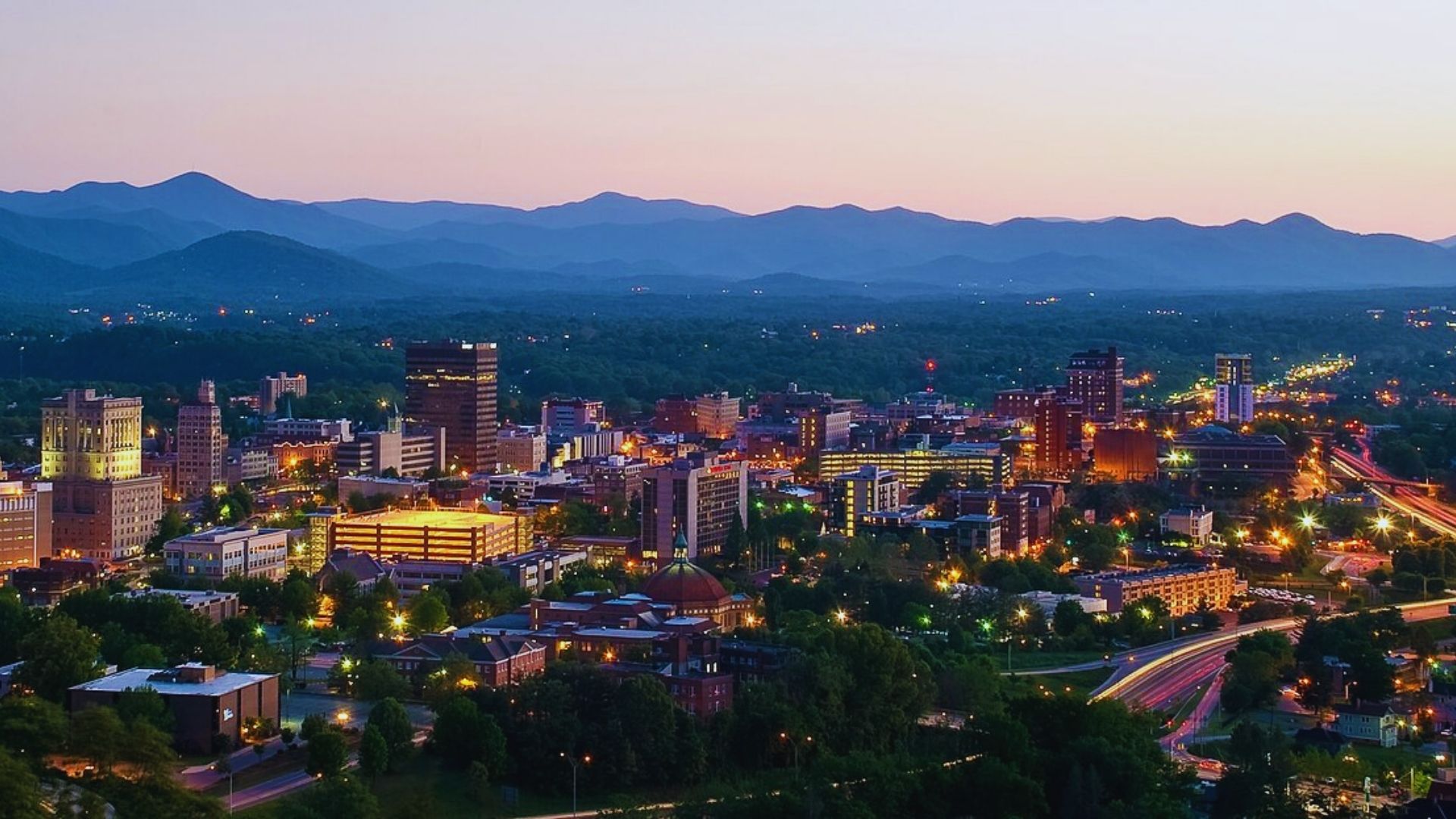 Asheville, NC skyline at dusk, showcasing illuminated buildings against a twilight sky.