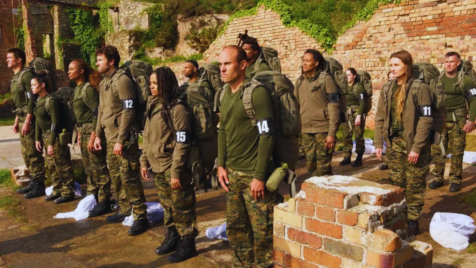A group of soldiers stands in formation in front of a weathered brick wall, dressed in military uniforms.