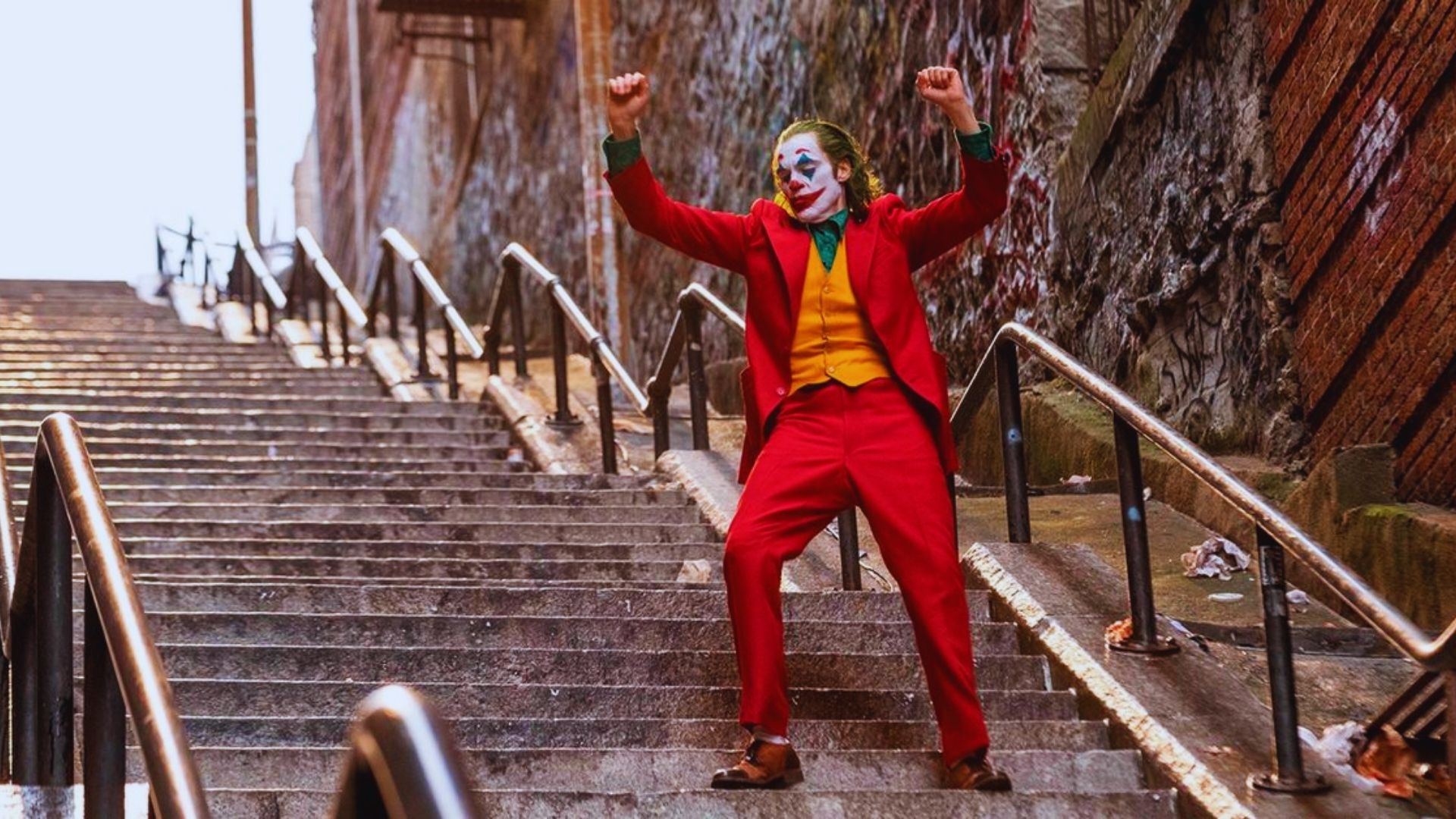 The Joker stands on the stairs of a building, dressed in his iconic purple suit and menacingly grinning at the camera.