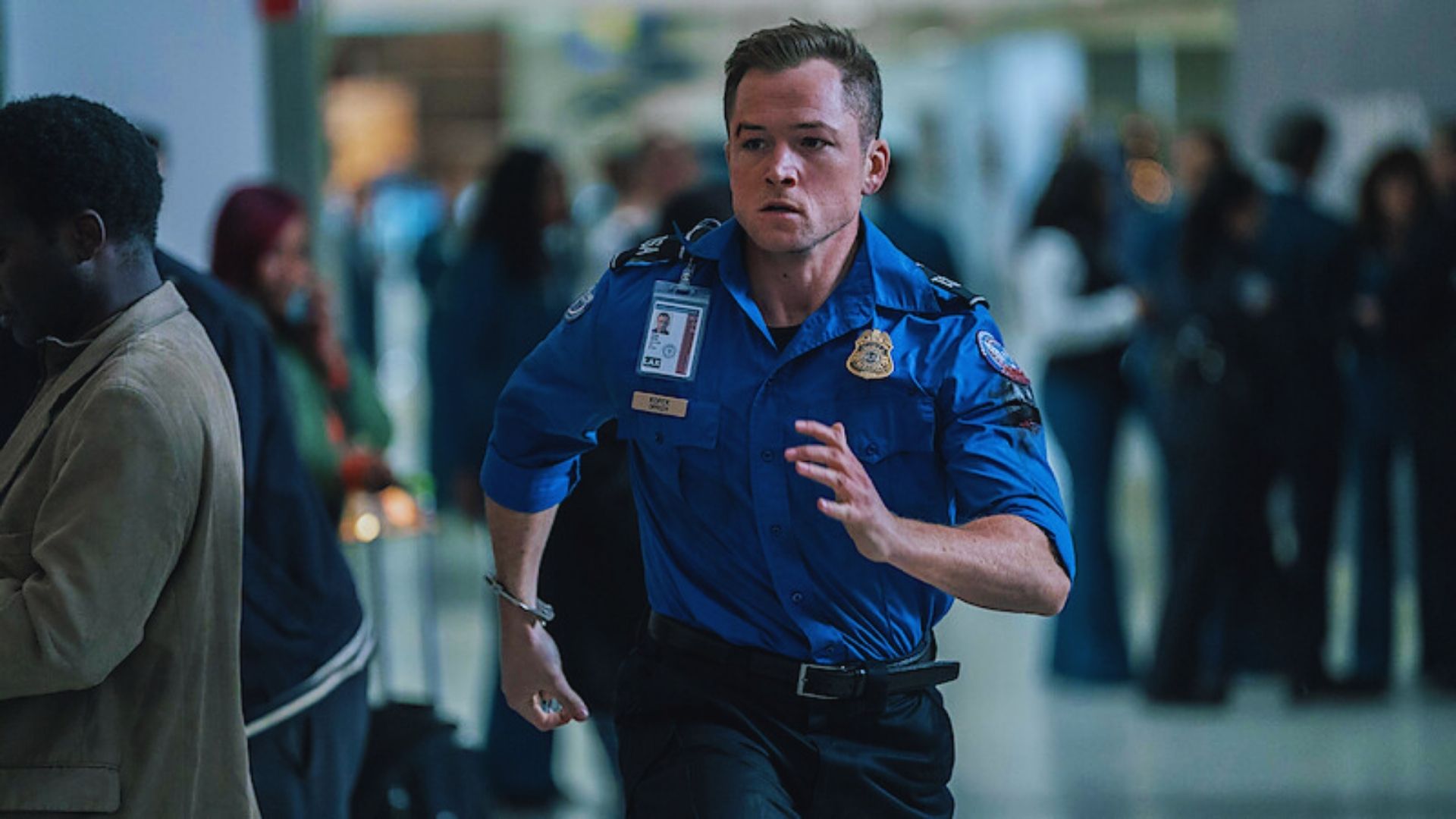 A man in a blue shirt is sprinting through an airport, surrounded by travelers and airport signage.

