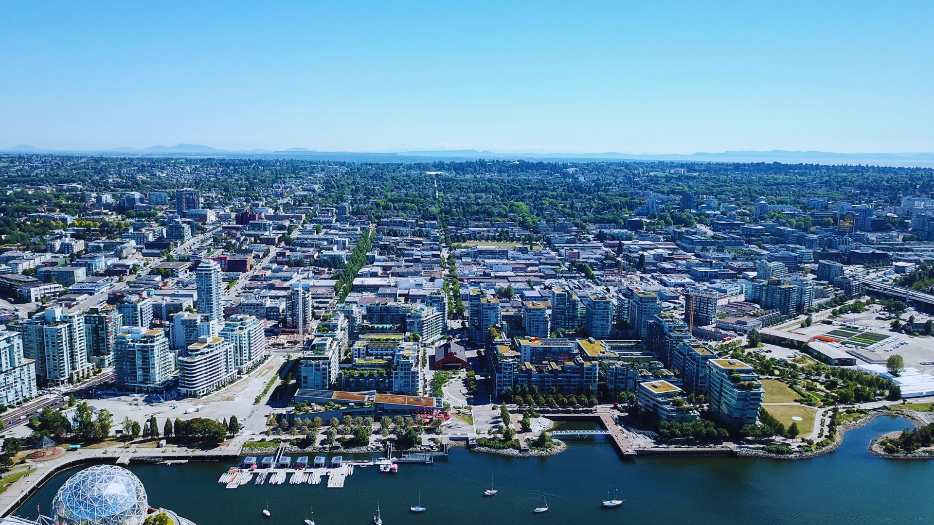 Aerial view of Vancouver showcasing its skyline, waterfront, and surrounding mountains under a clear blue sky.