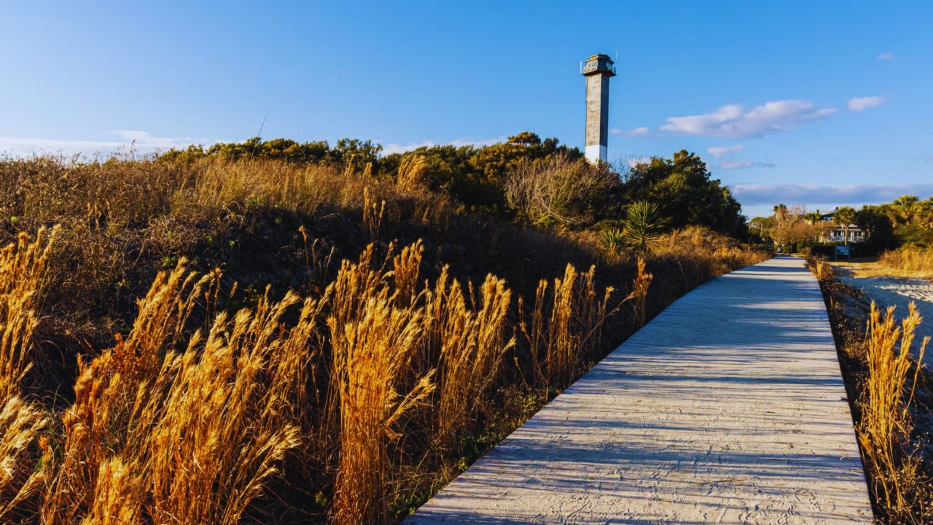 A wooden boardwalk stretches across the sand, leading to a tall lighthouse beside the beach under a clear blue sky.