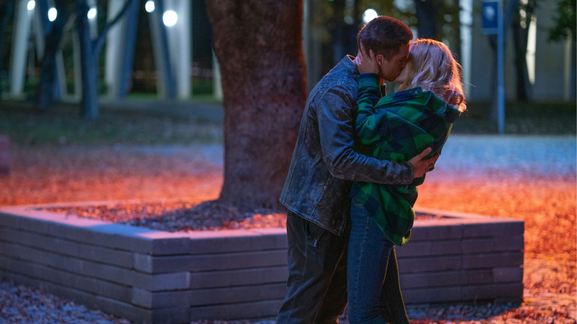 A young couple kisses tenderly in a park at night, with the soft glow of streetlights creating a romantic atmosphere.