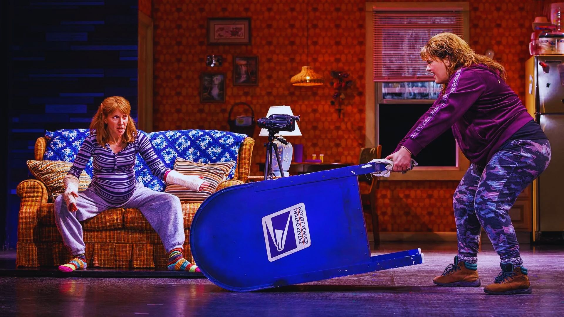Two women are in a staged living room. One sits on a couch, surprised, while the other struggles to lift a large blue USPS mailbox, creating a humorous scene.