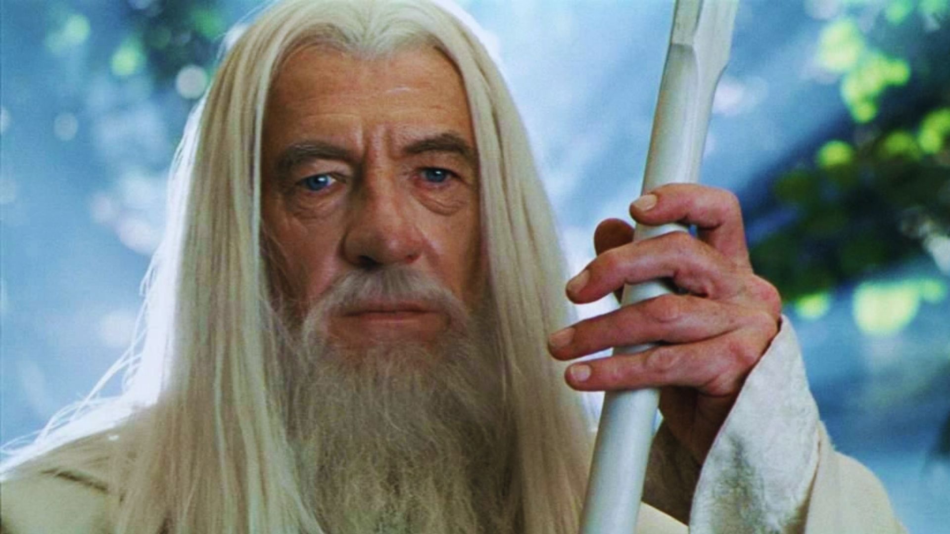 Elderly figure with long white hair and beard, holding a staff, stands in a serene forest setting. His expression is wise and thoughtful.