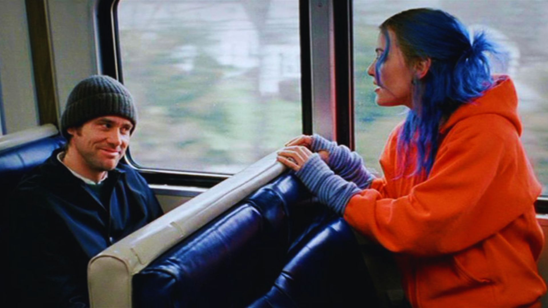 A person in a knit hat smiles at another with vibrant blue hair and an orange hoodie inside a train. The scene conveys a sense of connection and warmth.