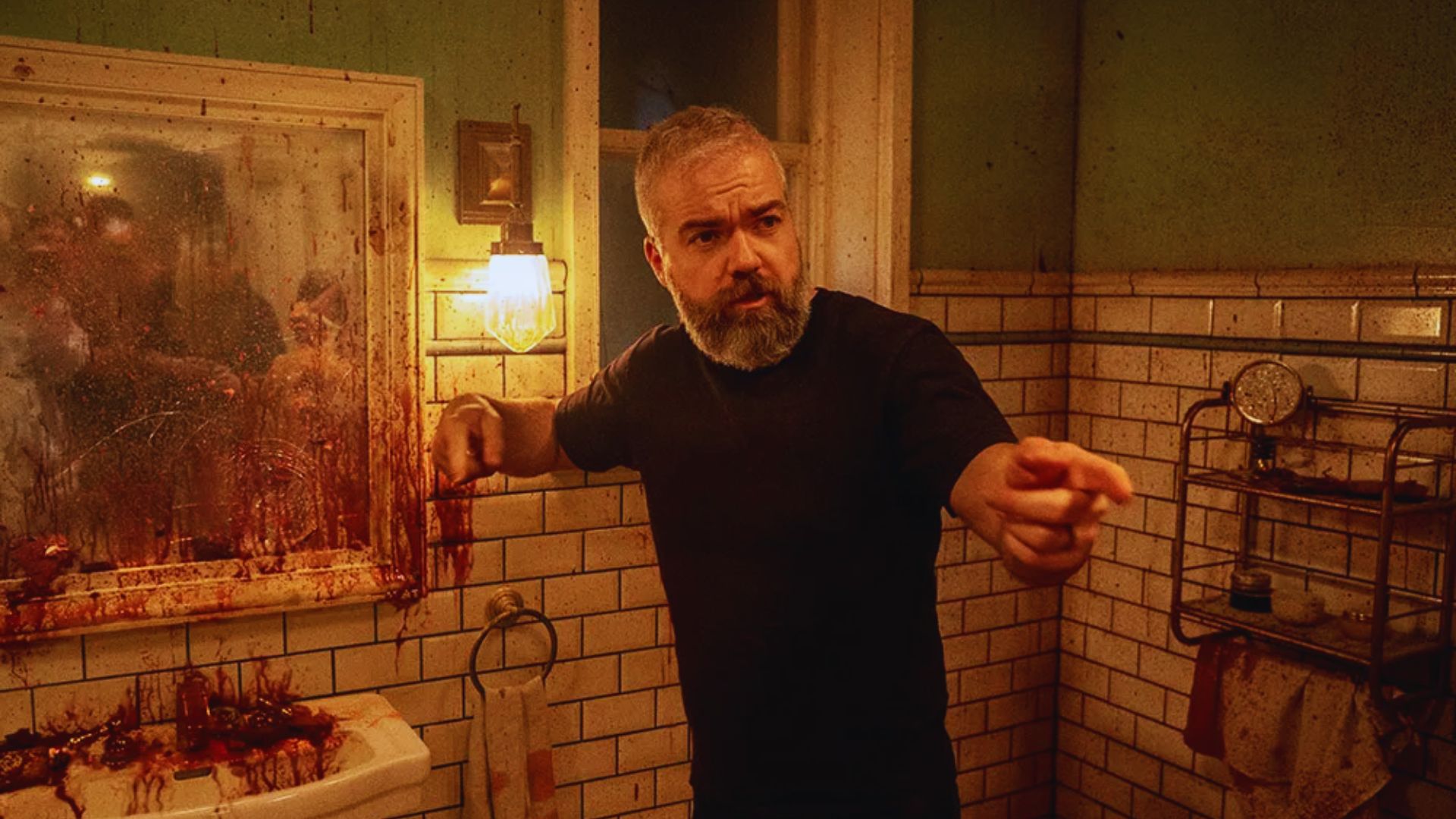 A man with a gray beard stands in a dimly lit, blood-splattered bathroom, pointing intensely. The walls are tiled, evoking a tense, eerie atmosphere.