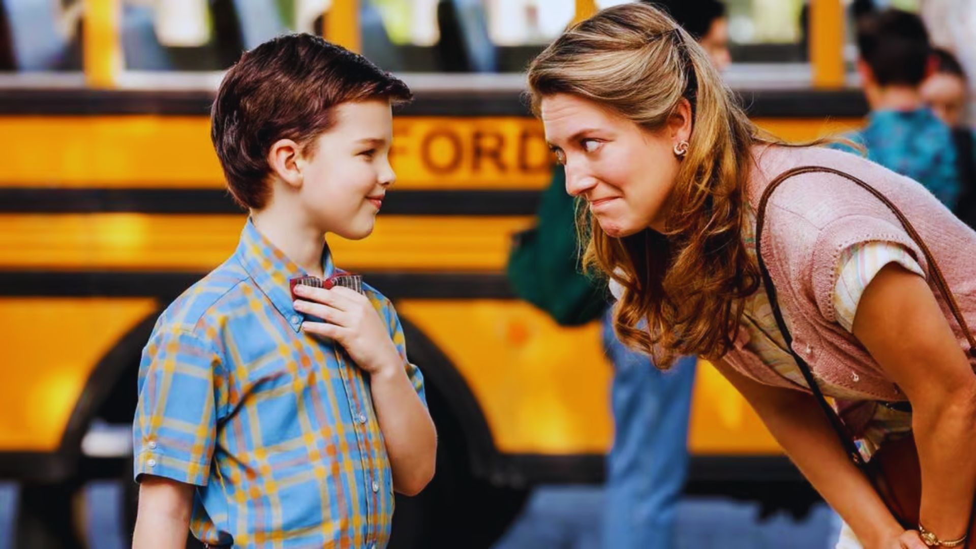 A smiling woman leans toward a young boy in a plaid shirt in front of a yellow school bus. They share a tender, affectionate moment.