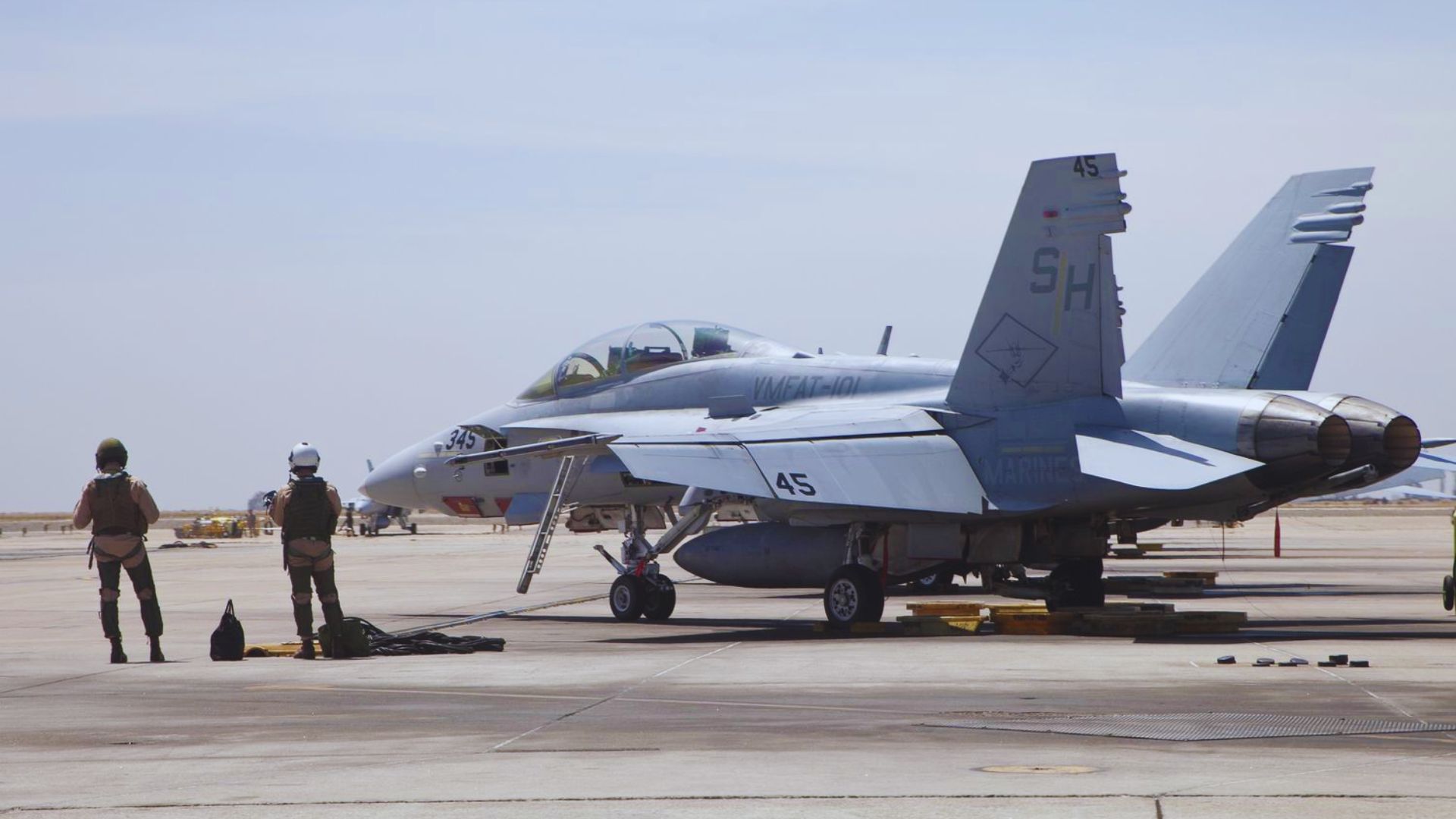 Two military personnel in flight gear stand on a runway near a parked F/A-18 Hornet jet. The scene is calm, with clear skies and open space around them.