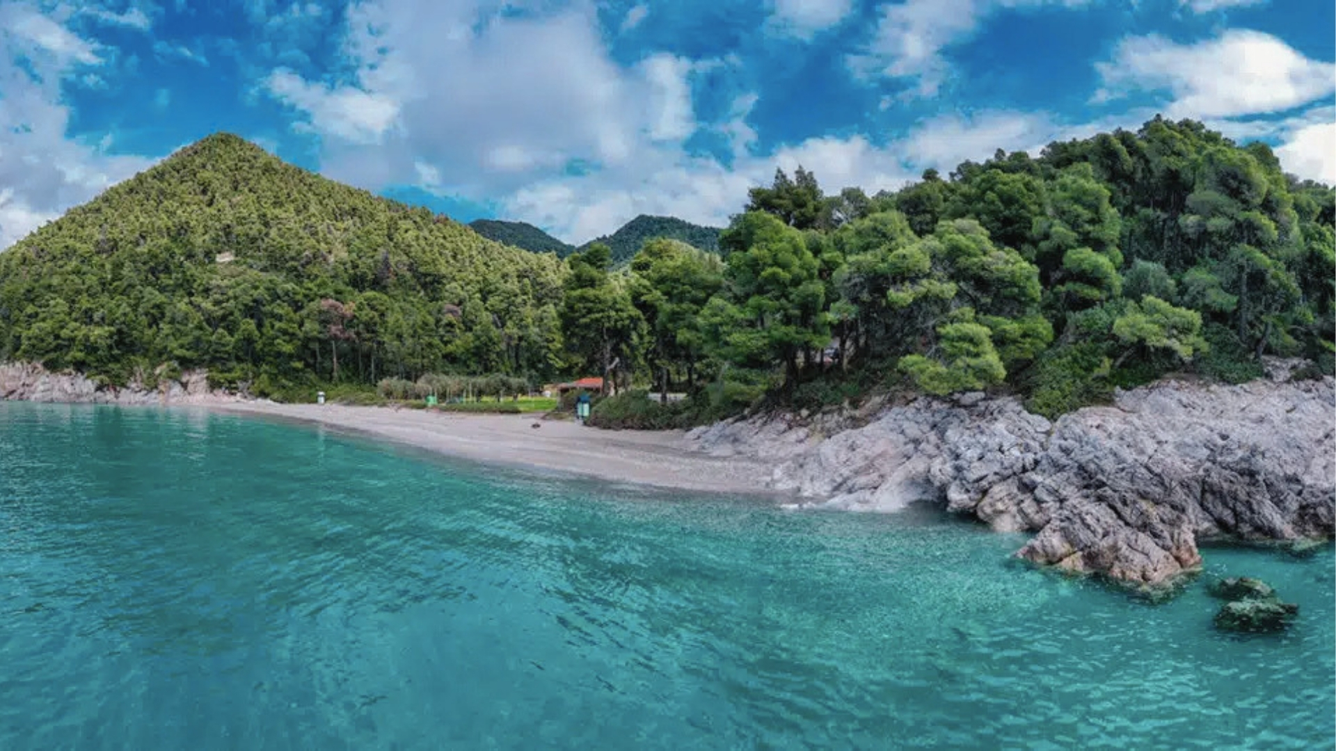 Small beach surrounded by lush forested hills, turquoise water, sandy shore, rocky outcrops, and bright sky in Greece.

