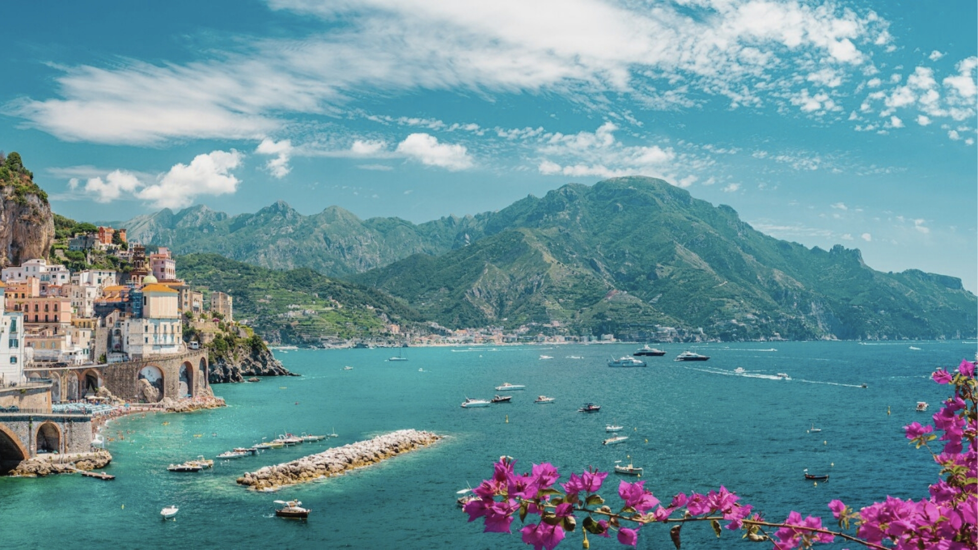 Colorful Amalfi Coast village cascading down cliffs to turquoise bay, boats, pink flowers, Hotel Costiera filming location.
