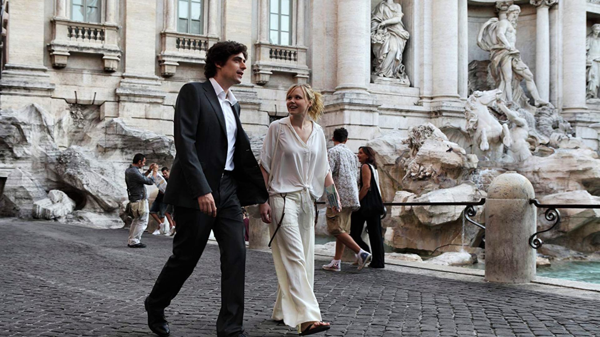 A man and woman stroll together in front of a beautiful fountain, capturing a romantic moment in Rome.