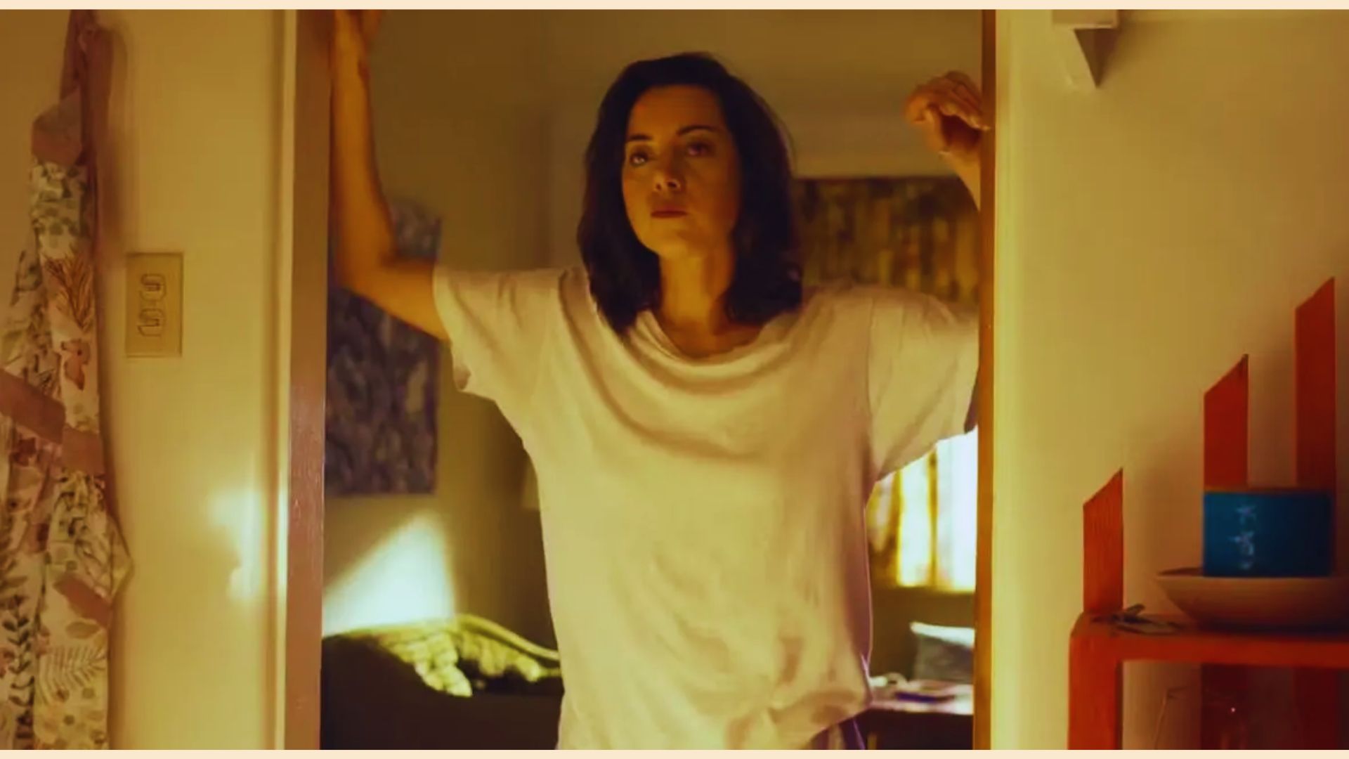 Woman in a relaxed white t-shirt stands in a warmly lit doorway, arms up, with a thoughtful expression. Soft interior colors convey a cozy mood.
