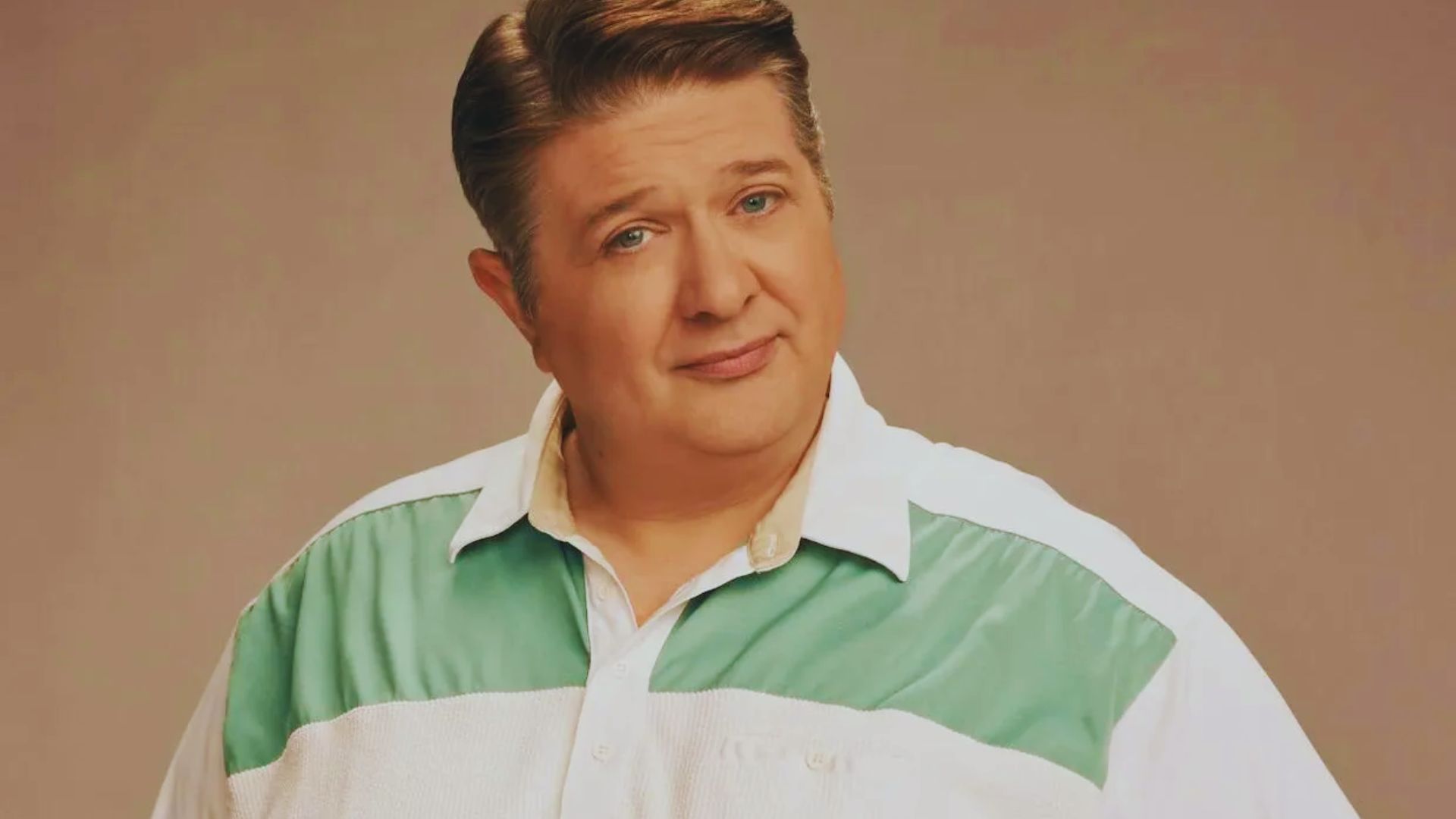 A man with short, neatly styled hair and a relaxed smile wears a button-up shirt with green and white panels. The background is a neutral shade.