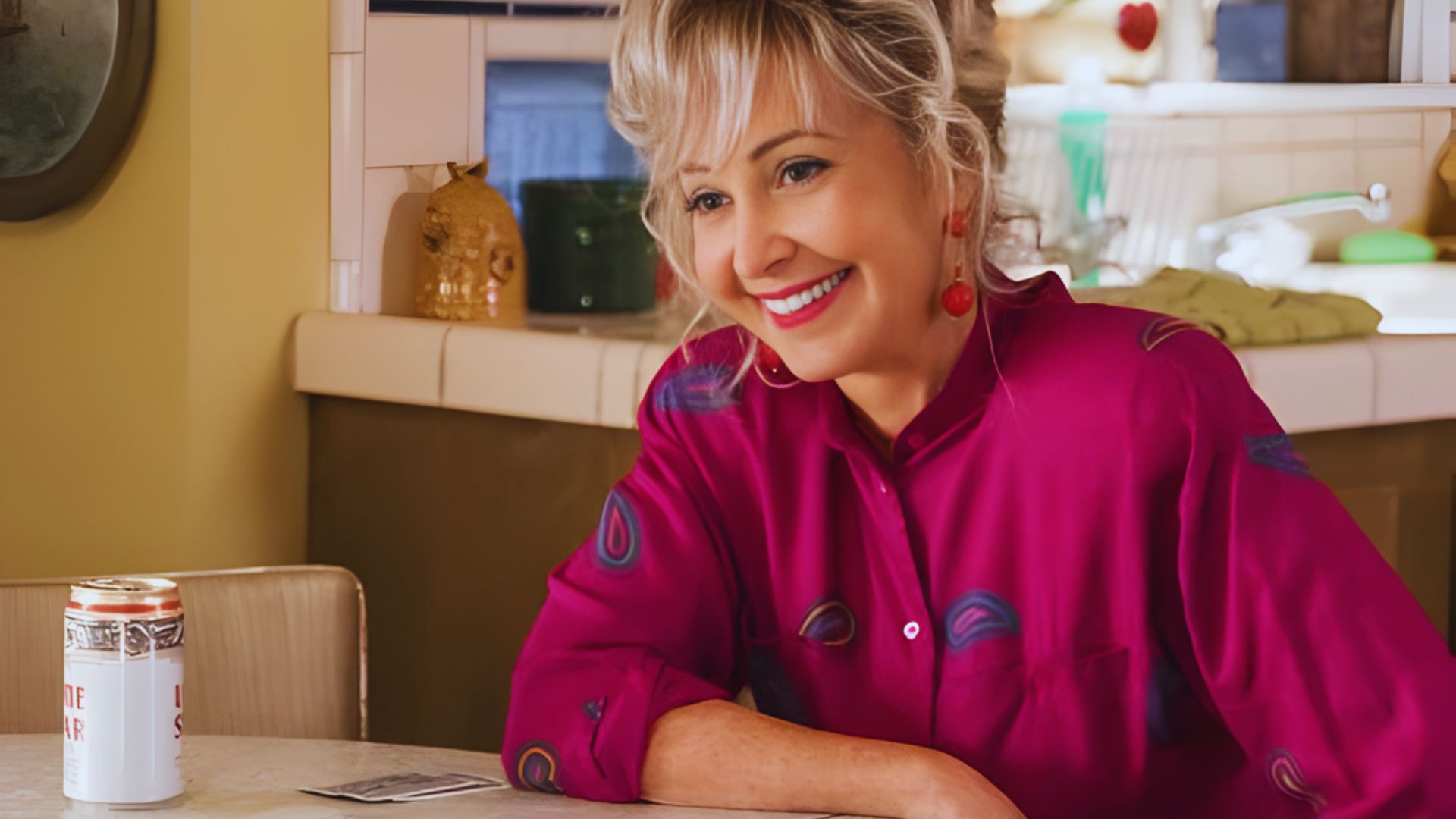 Smiling woman in a pink blouse with a pattern sits at a kitchen table. A can and cards are in front of her, adding a casual, cheerful vibe.
