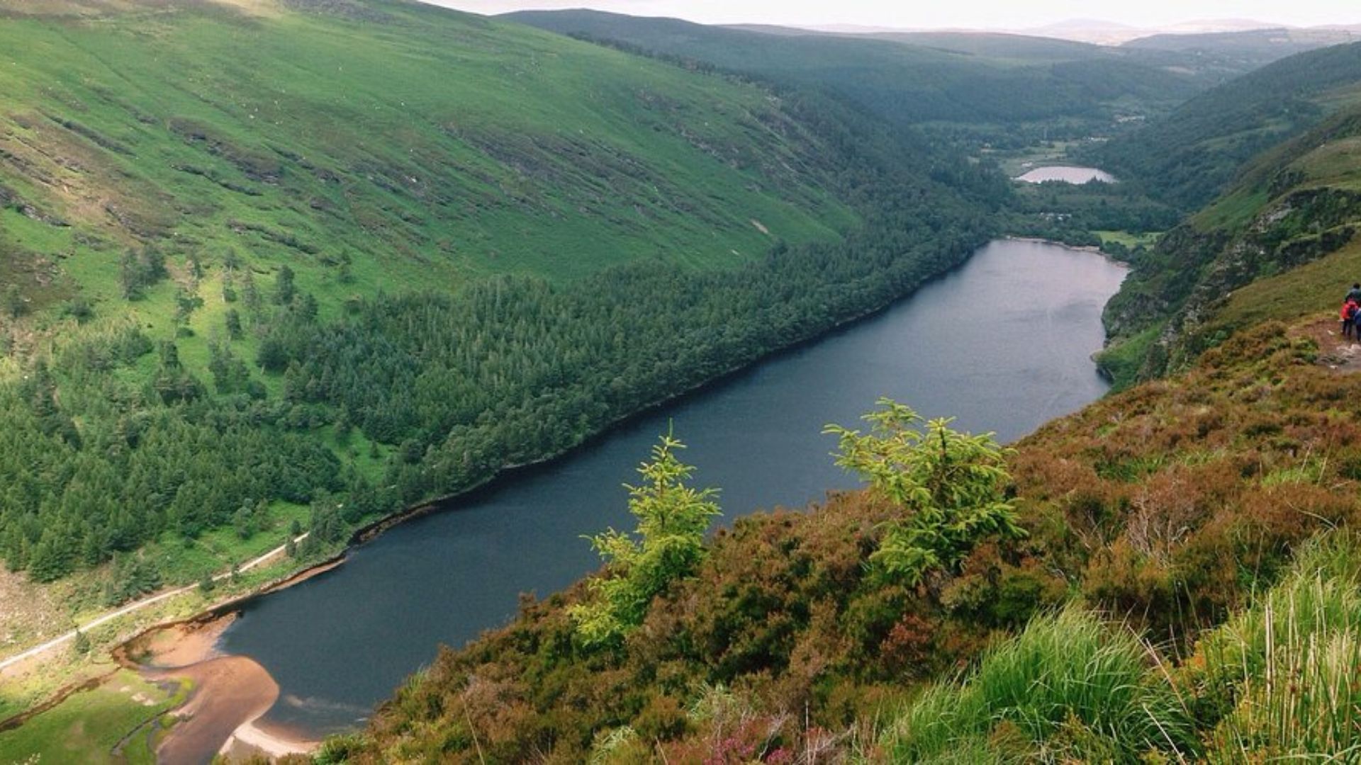 A serene landscape of a lush, green valley surrounding a calm lake. The scene conveys tranquility, with gentle hills and dense forested areas.