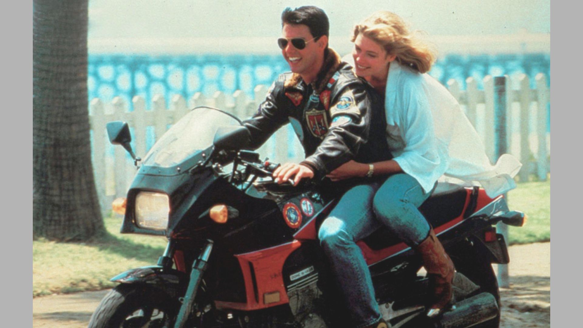 Man and woman ride a motorcycle on a sunny day. He wears a dark jacket and sunglasses; she holds him, smiling, with wind in her hair. Fence and trees are in the background.