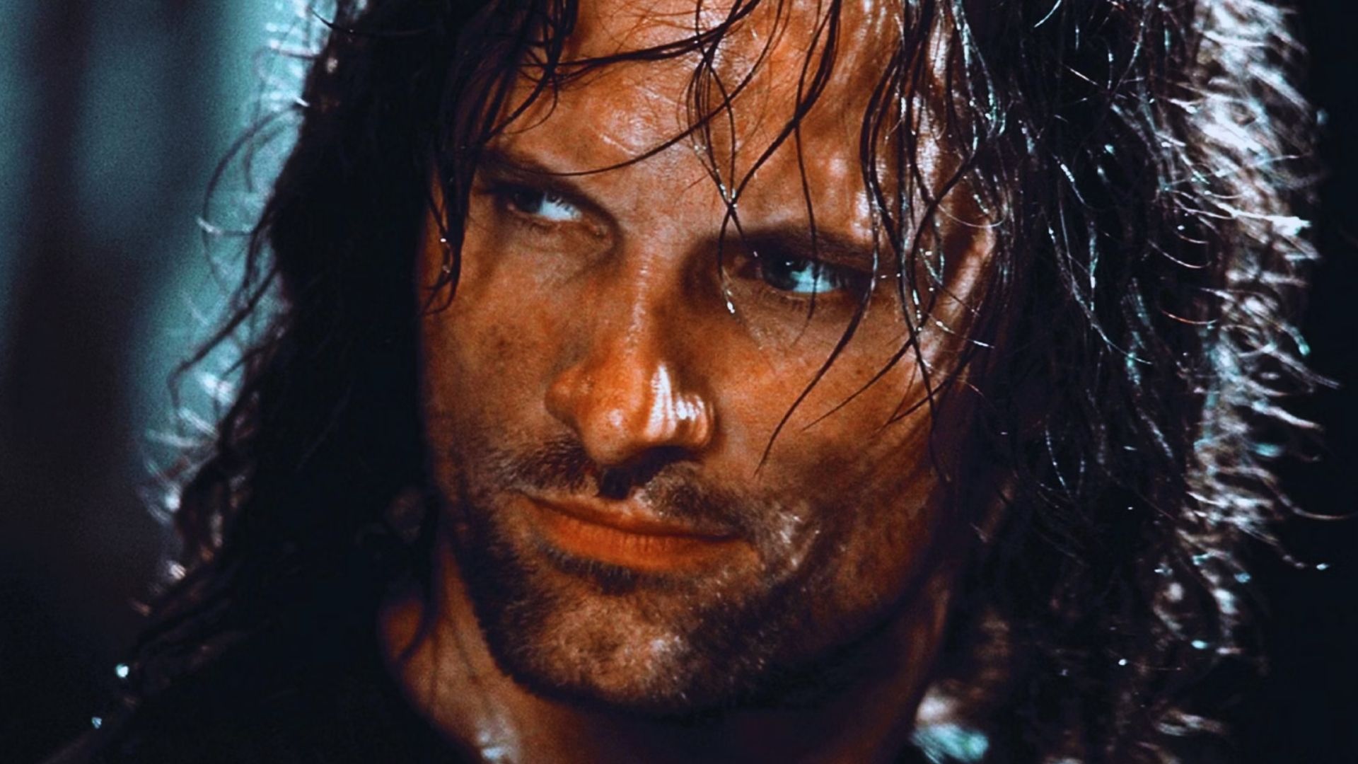 A rugged man with wet, shoulder-length hair and intense blue eyes stares to the side. His face is dirty, conveying a determined and fierce expression.