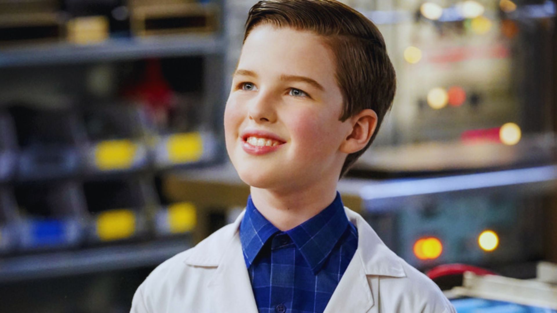 A young boy with a short haircut smiles confidently, wearing a white lab coat over a blue checkered shirt. Blurred lab equipment is visible in the background.