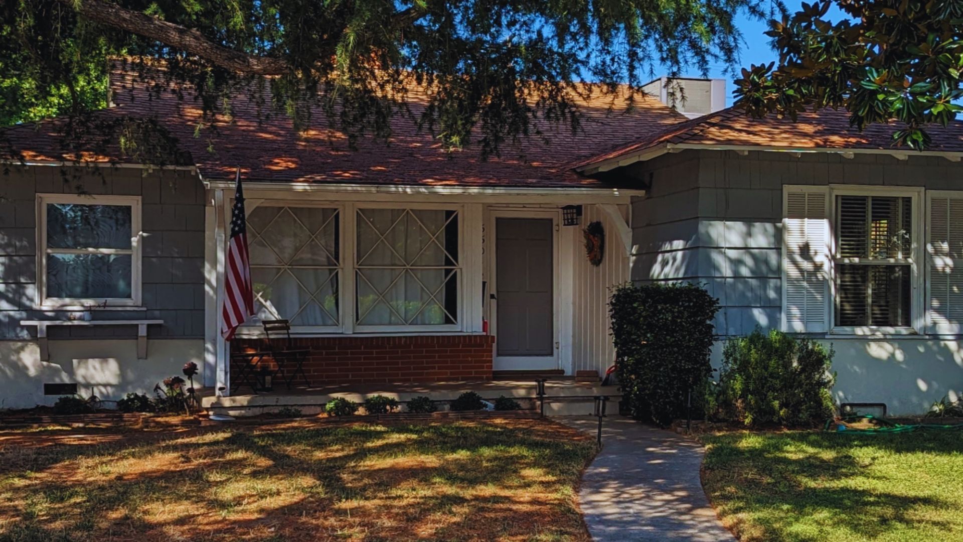 A quaint suburban house with gray siding and a brown roof, fronted by a small porch. An American flag is displayed, with greenery and trees around.