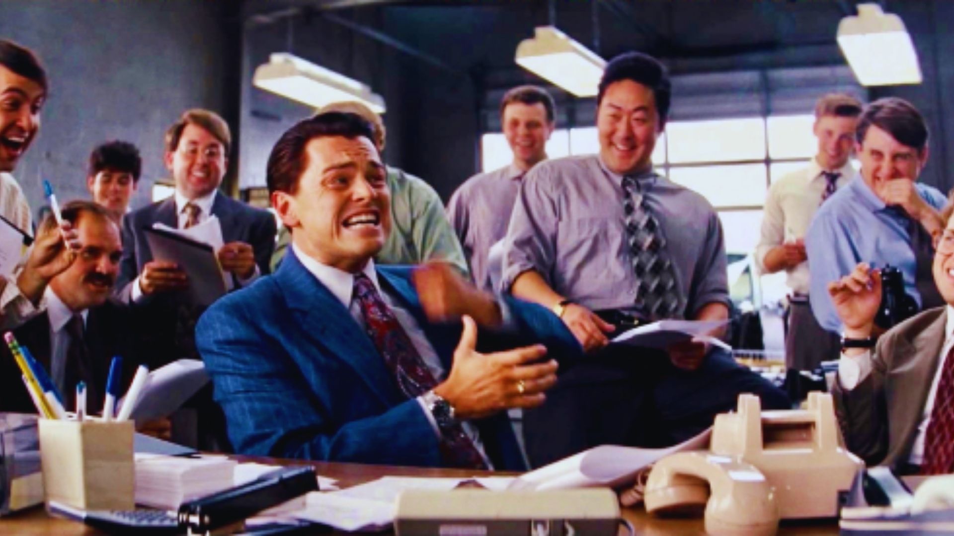 A lively office scene with a group of men in shirts and ties, joyfully laughing around a desk cluttered with papers and a phone, conveying a humorous, energetic atmosphere.