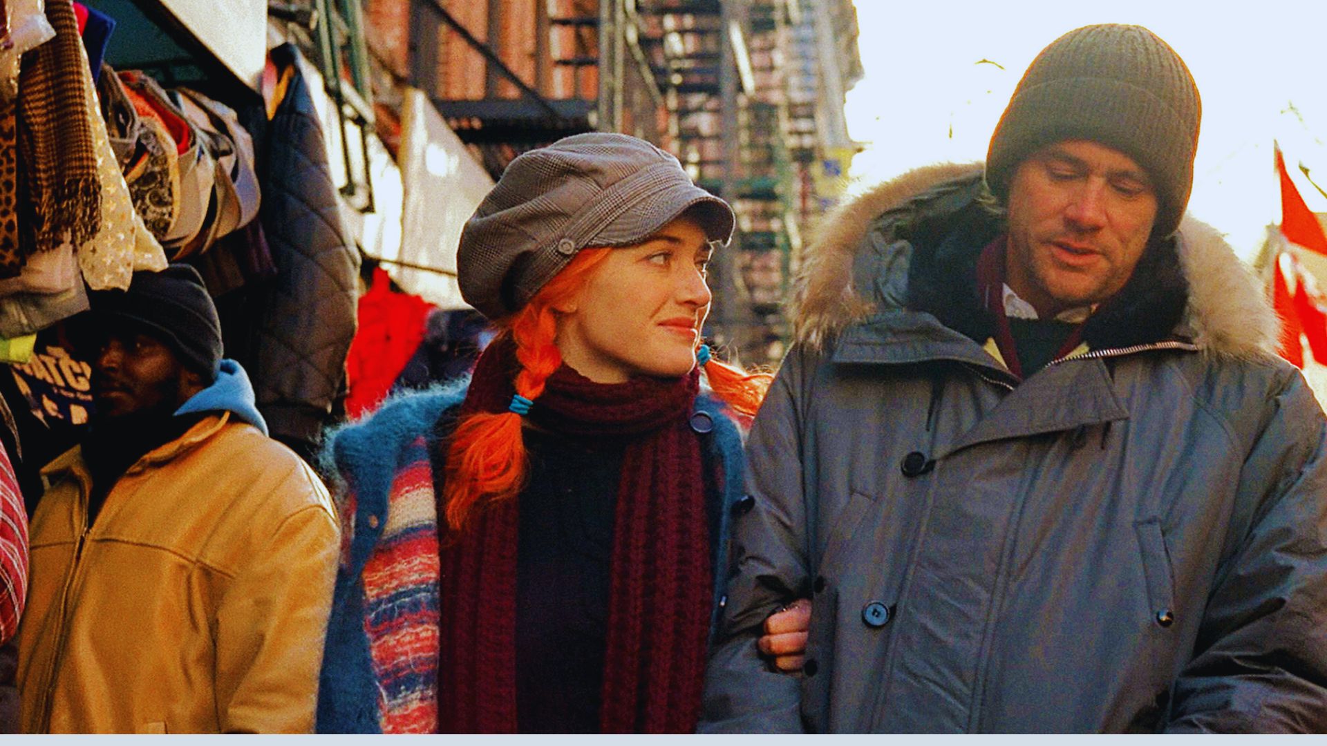A woman with bright red hair and a gray cap smiles at a man in a winter coat as they walk arm-in-arm through a bustling, warm-toned street market.