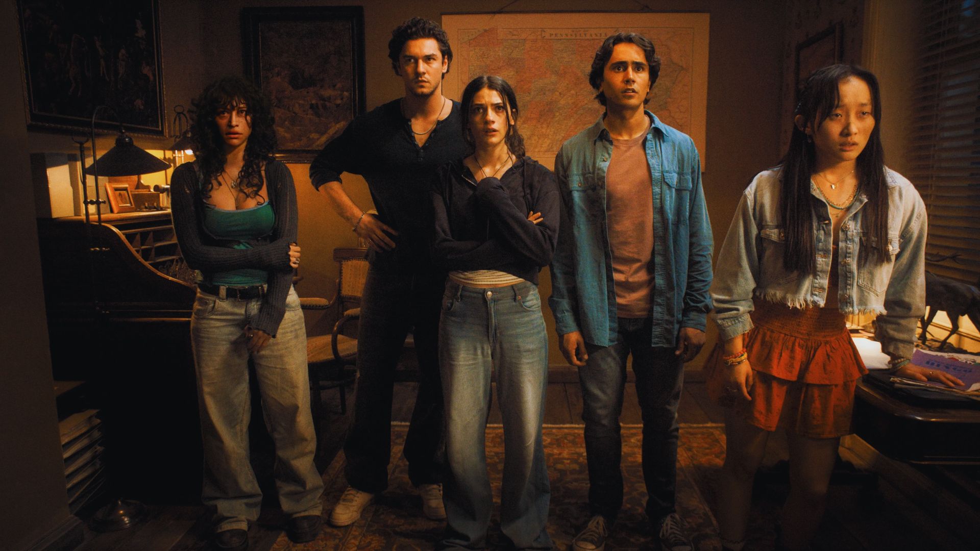 Five young adults stand closely in a dim room with serious expressions. A world map and a desk lamp illuminate the cozy, mysterious setting.