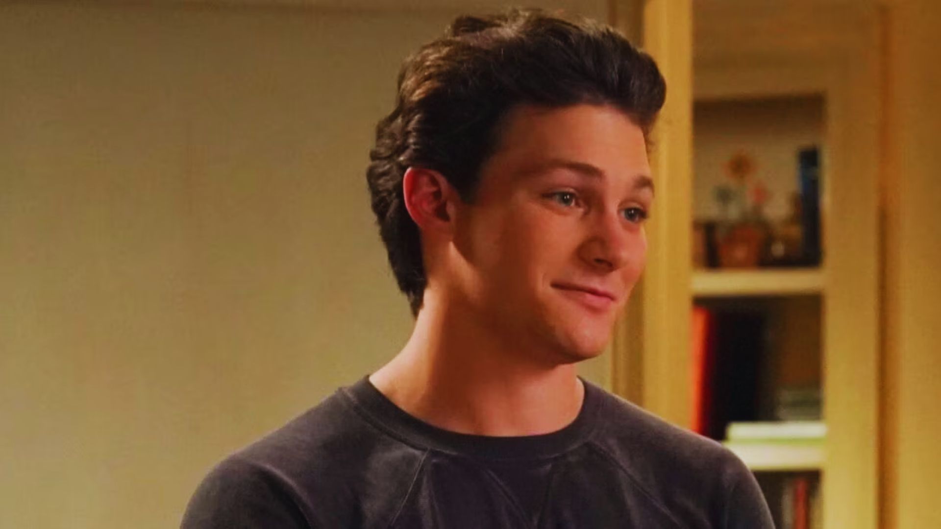 Young man with short dark hair smiling softly, wearing a dark sweater, standing indoors near shelves with books and plants. Warm, casual atmosphere.