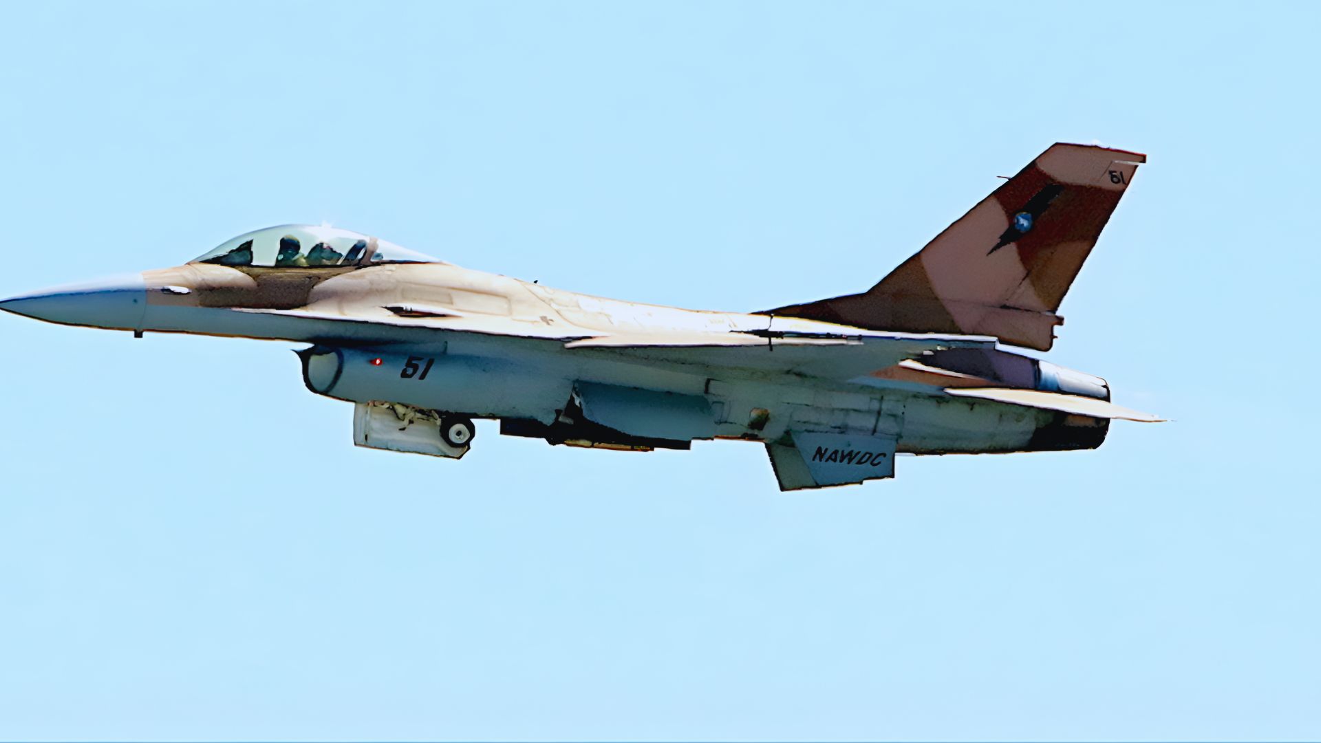 Fighter jet in flight with a desert camouflage pattern against a clear blue sky. The cockpit windows are visible, conveying speed and precision.