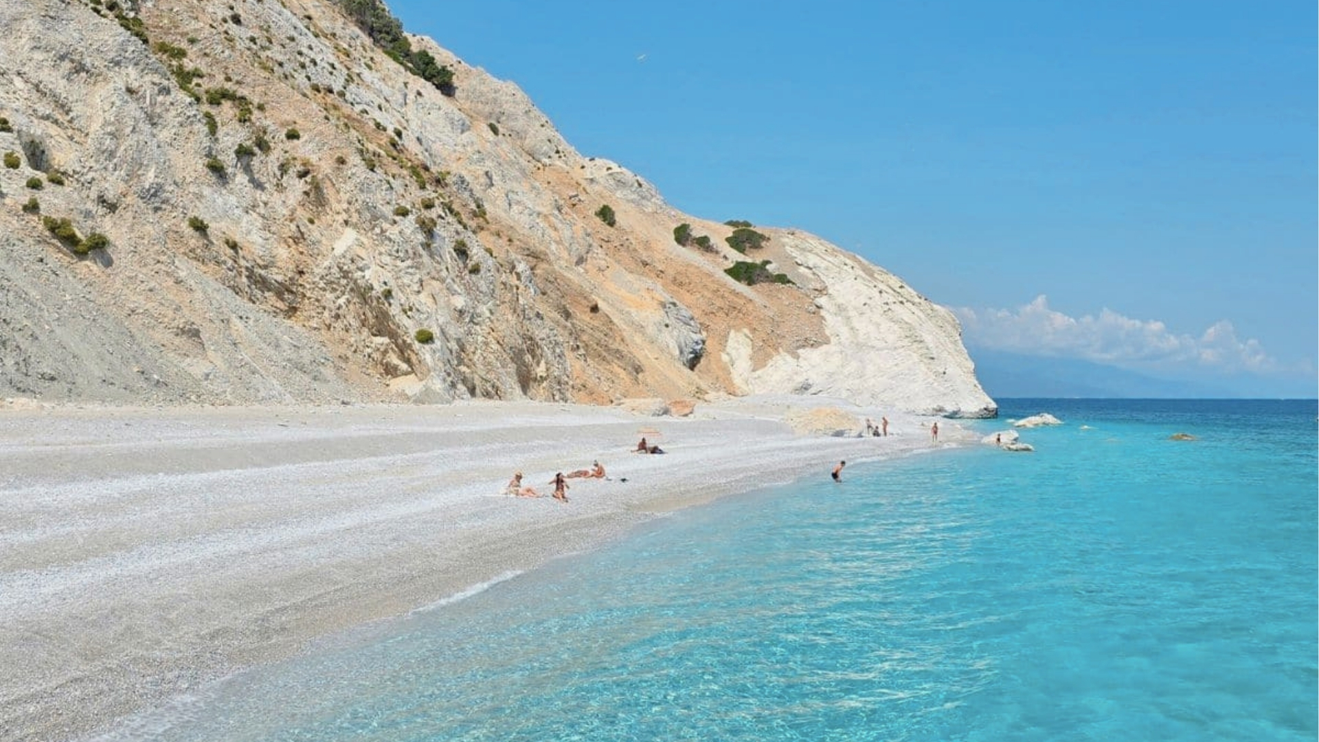 Rocky cliff with sparse vegetation, pebbly beach, turquoise water, bright sky, people swimming, and relaxing in Greece.