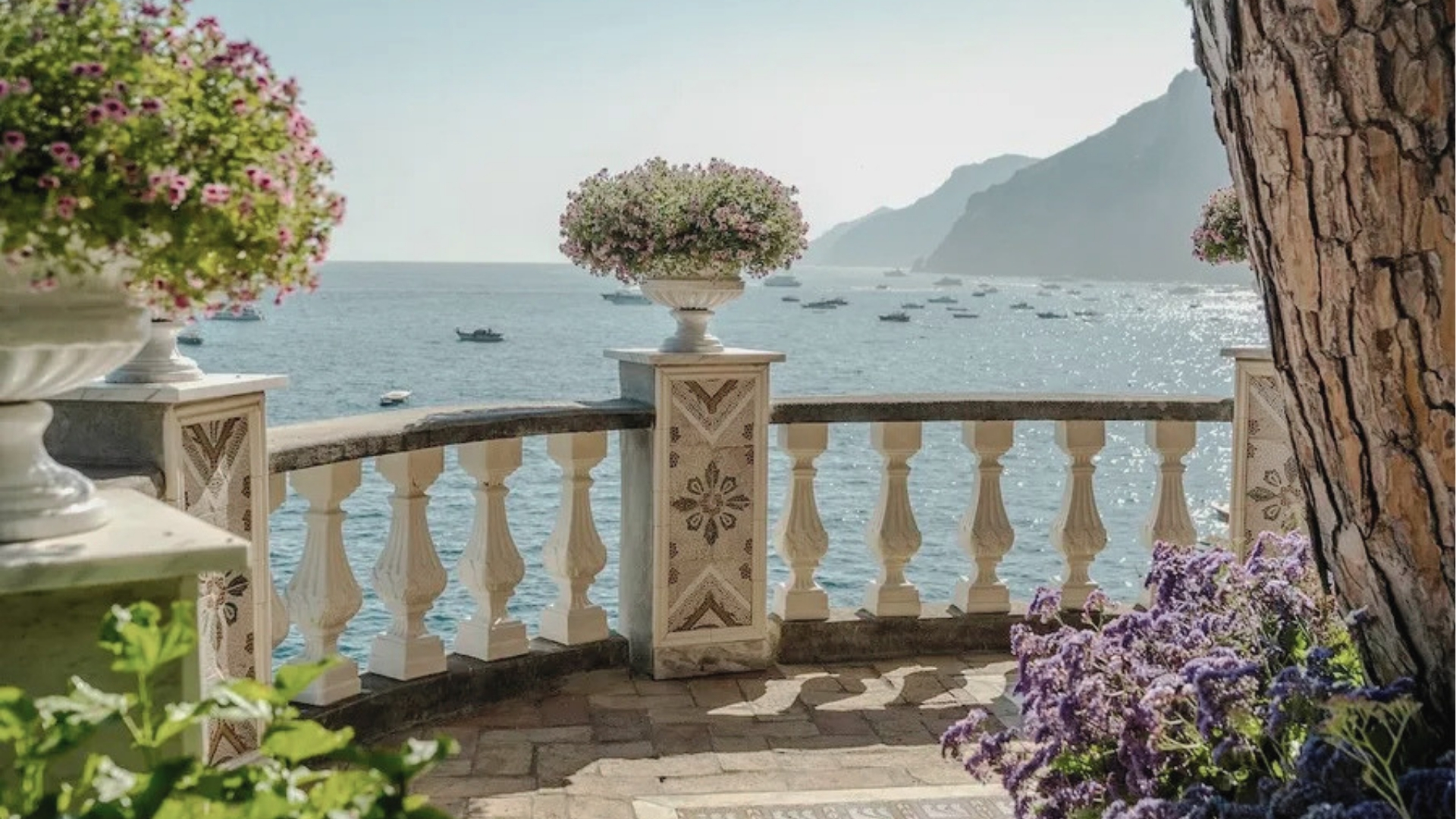 Sunlit stone terrace with mosaic railings, flower urns, boats on sparkling sea, mountains, Hotel Costiera filming location.
