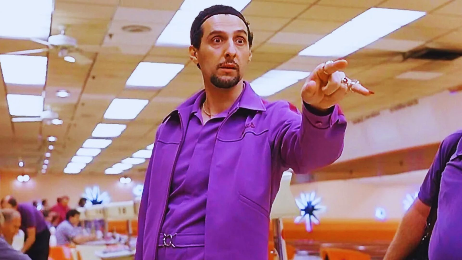 A man in a purple jumpsuit dramatically points forward in a brightly lit bowling alley. The atmosphere is intense and focused, highlighting his confident and assertive pose.