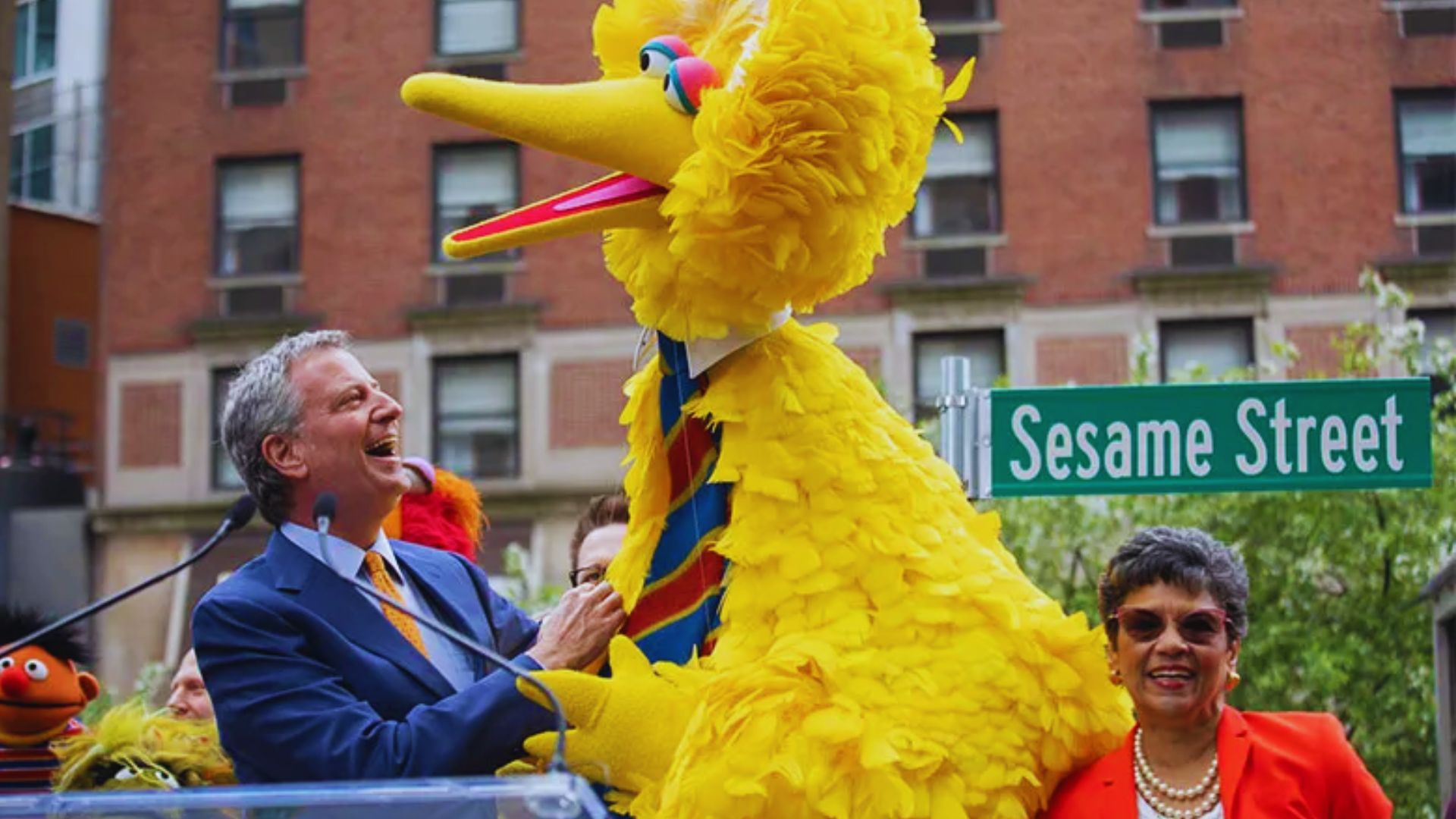 Big Bird from Sesame Street with a man at a podium near a Sesame Street sign, surrounded by people in an outdoor city setting.