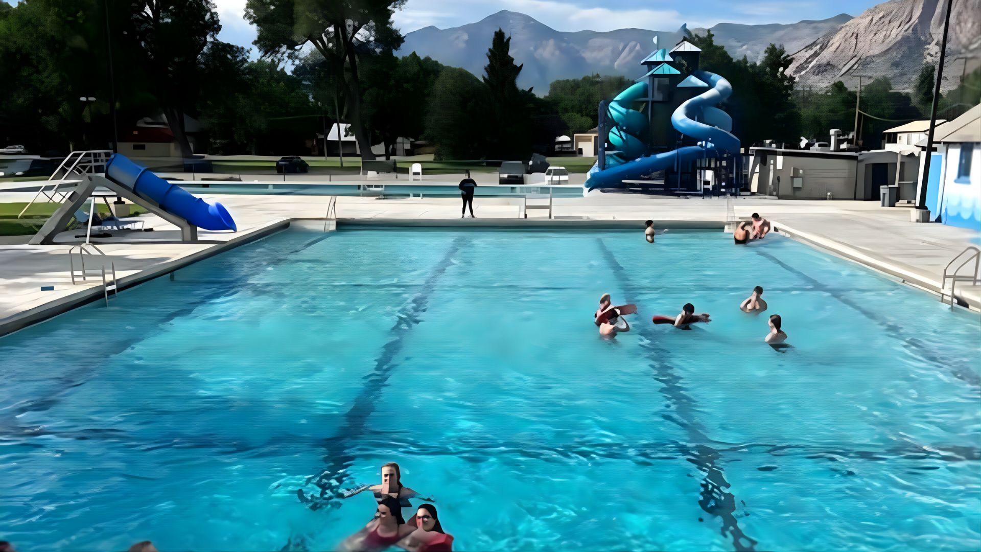 People swim in a pool at Lorin Farr Park, with scenic mountains visible in the background.
