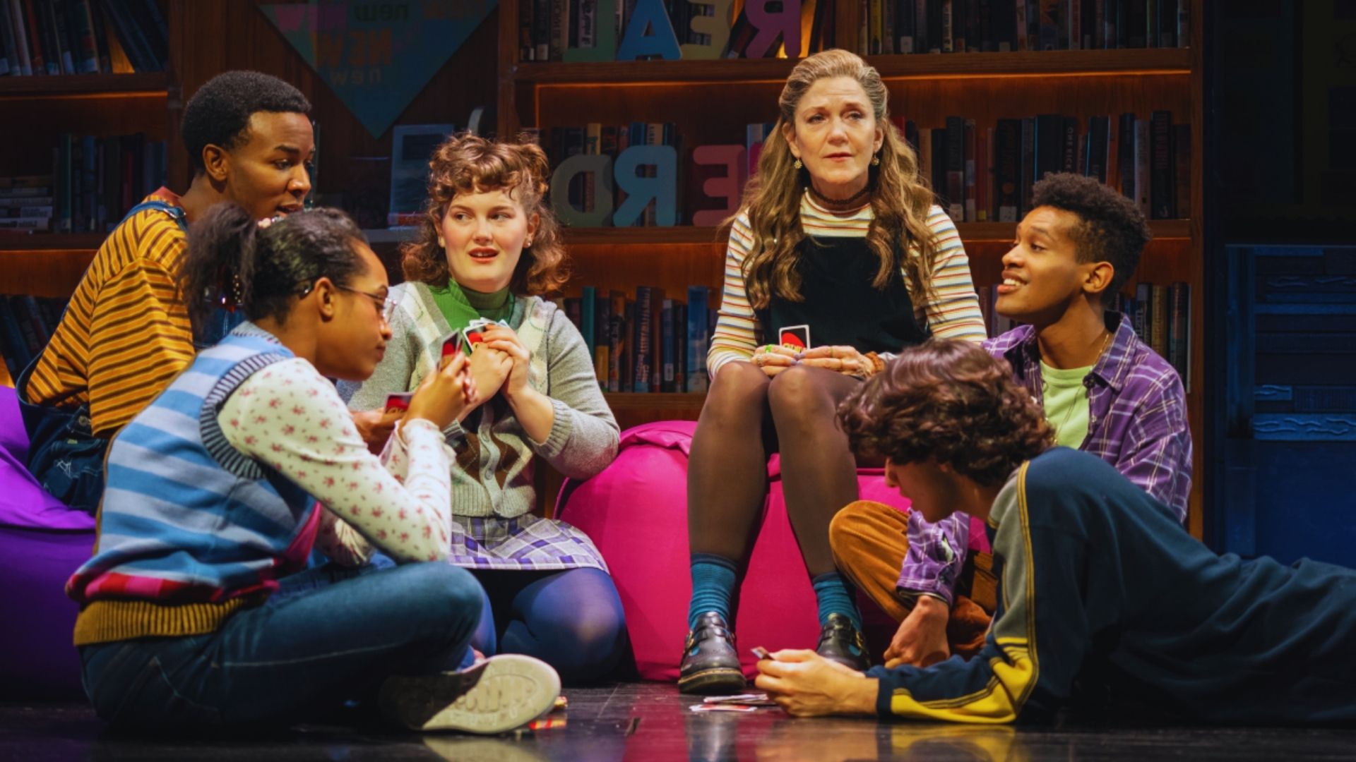 A group of six people sit on a vibrant purple couch in a library, engaging playfully with vibrant expressions. They are focused on a game of cards.