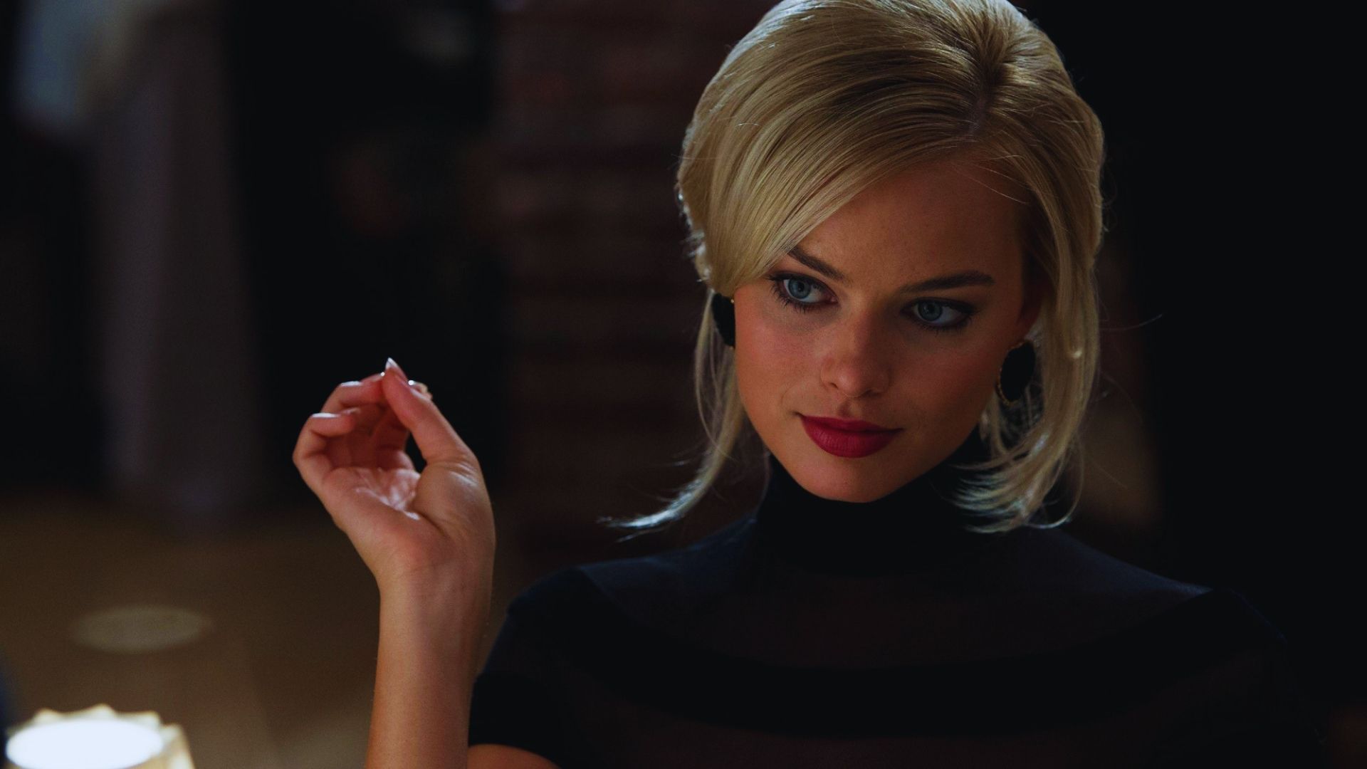 A woman with blonde hair and red lipstick gazes thoughtfully to her side in a dimly lit setting. She wears a black top, exuding elegance and intrigue.