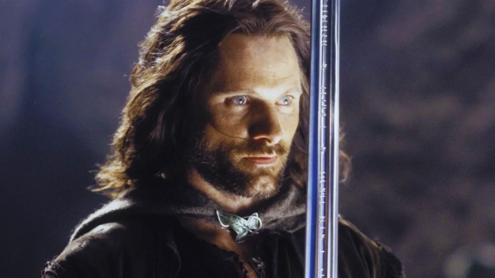 A man with long brown hair and a focused expression holds a gleaming sword vertically. He wears a cloak, evoking a mysterious, medieval atmosphere.