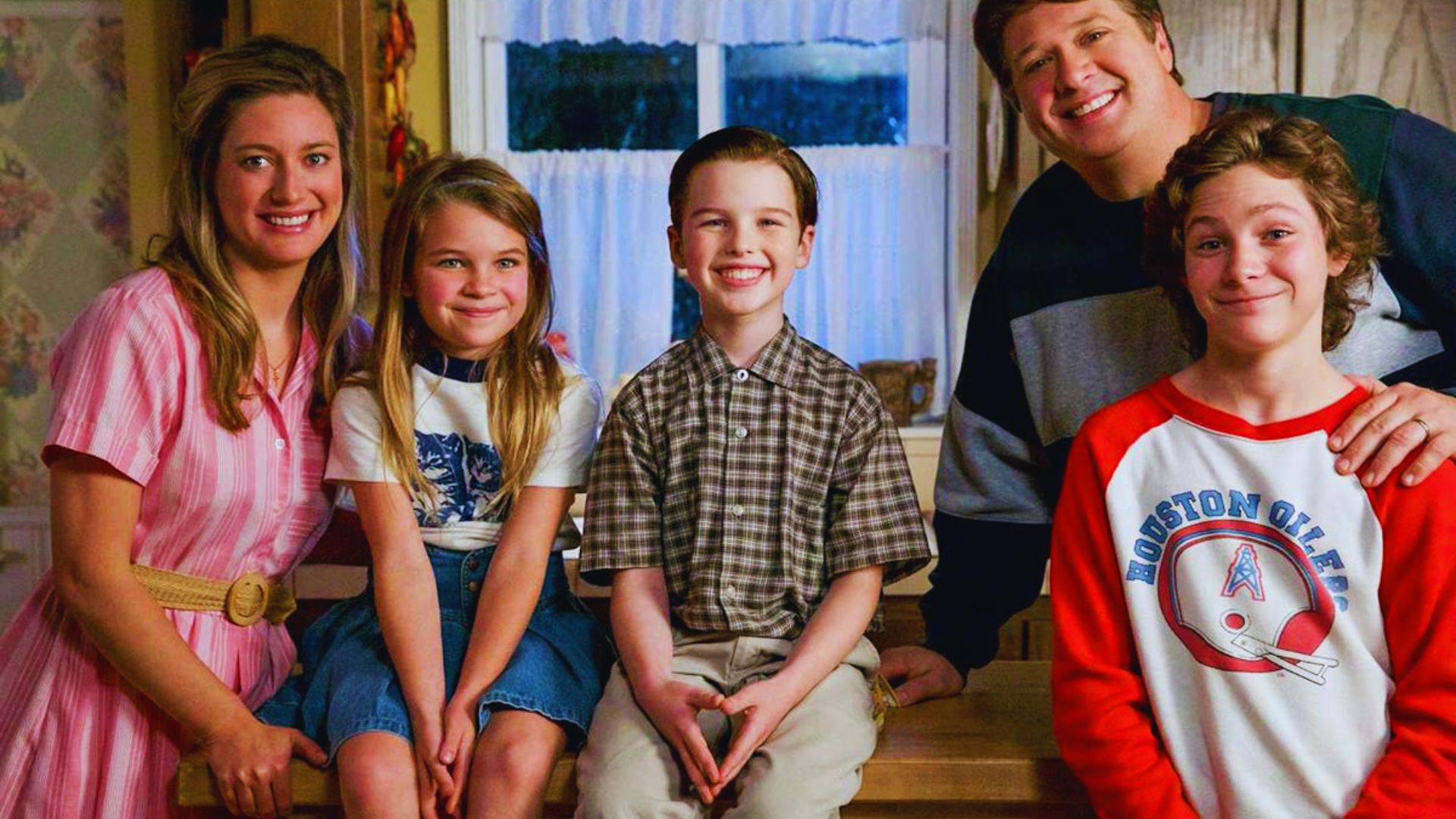 A family of five smiles warmly in a cozy kitchen. The children sit on a counter, the parents stand beside them. The setting feels joyful and inviting.