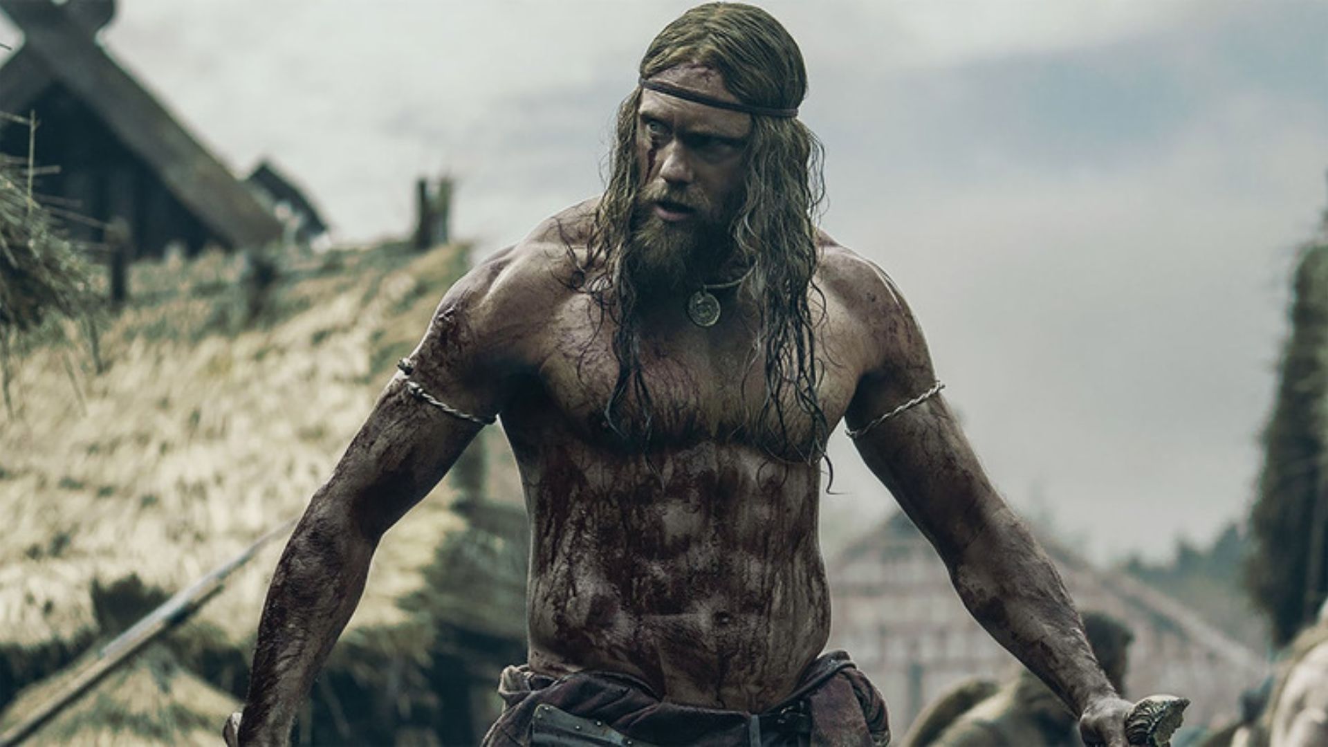 A muscular, bearded warrior stands fiercely, covered in dirt and blood. He's outdoors, with thatched huts in the background under a cloudy sky, evoking tension.