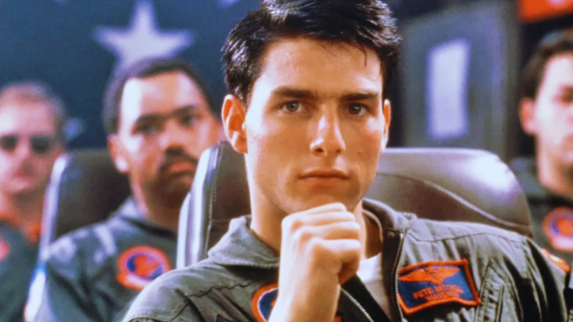 A man in a military uniform sits thoughtfully in a cockpit, with a hand resting near his face. Blurry figures and star insignias are visible in the background.