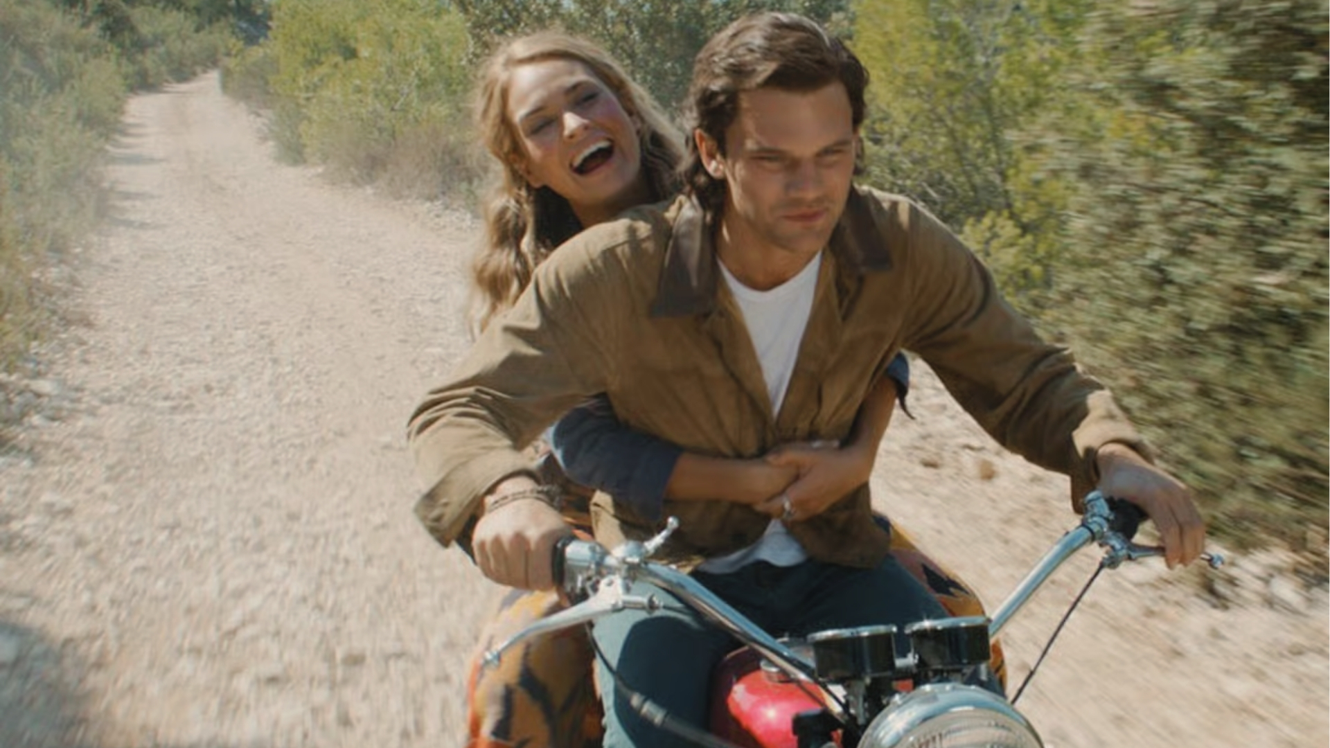 Two people on a motorcycle on a dirt road, surrounded by greenery, bushes, trees, and the countryside atmosphere in Greece.
