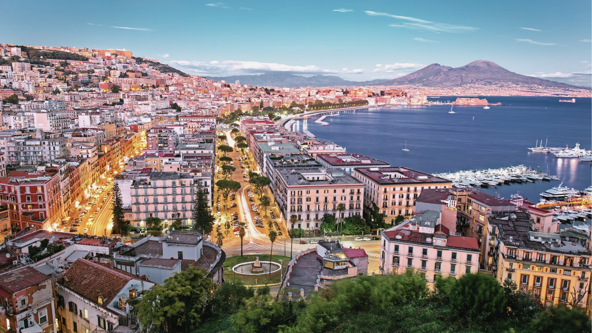 Aerial view of Naples waterfront, Vesuvius volcano, marina, city lights, palm trees, Hotel Costiera filming location.
