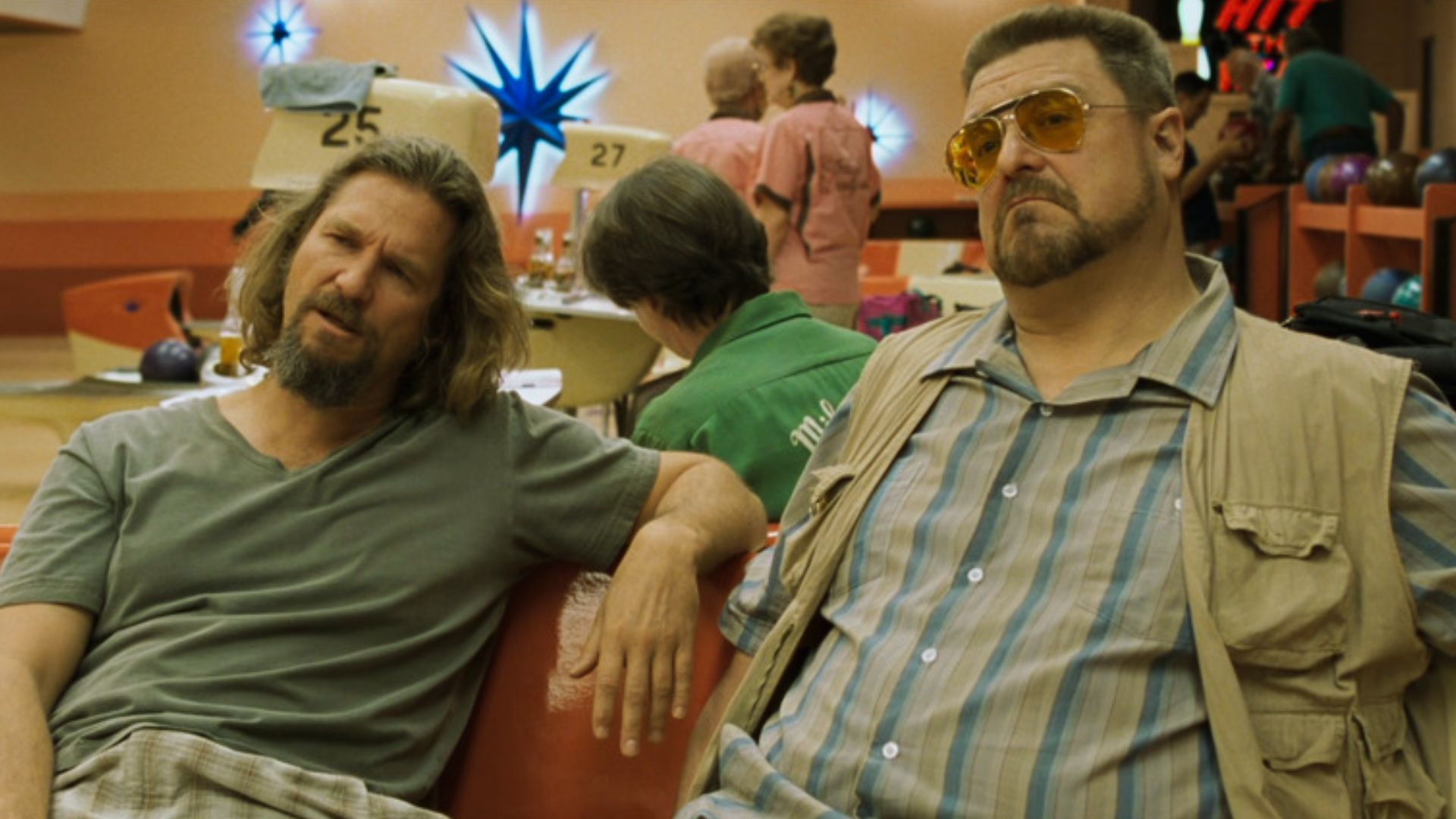 Two men sit on a red bench in a retro bowling alley. One wears a gray t-shirt, the other a vest with sunglasses. The scene feels laid-back and casual.