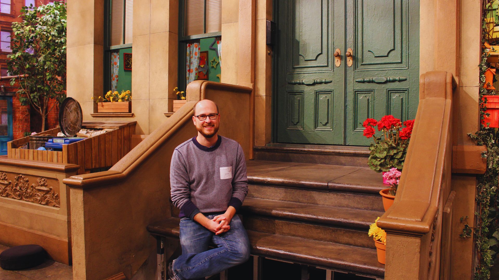 Man standing in front of a brown building surrounded by colorful plants in a scenic outdoor setting.